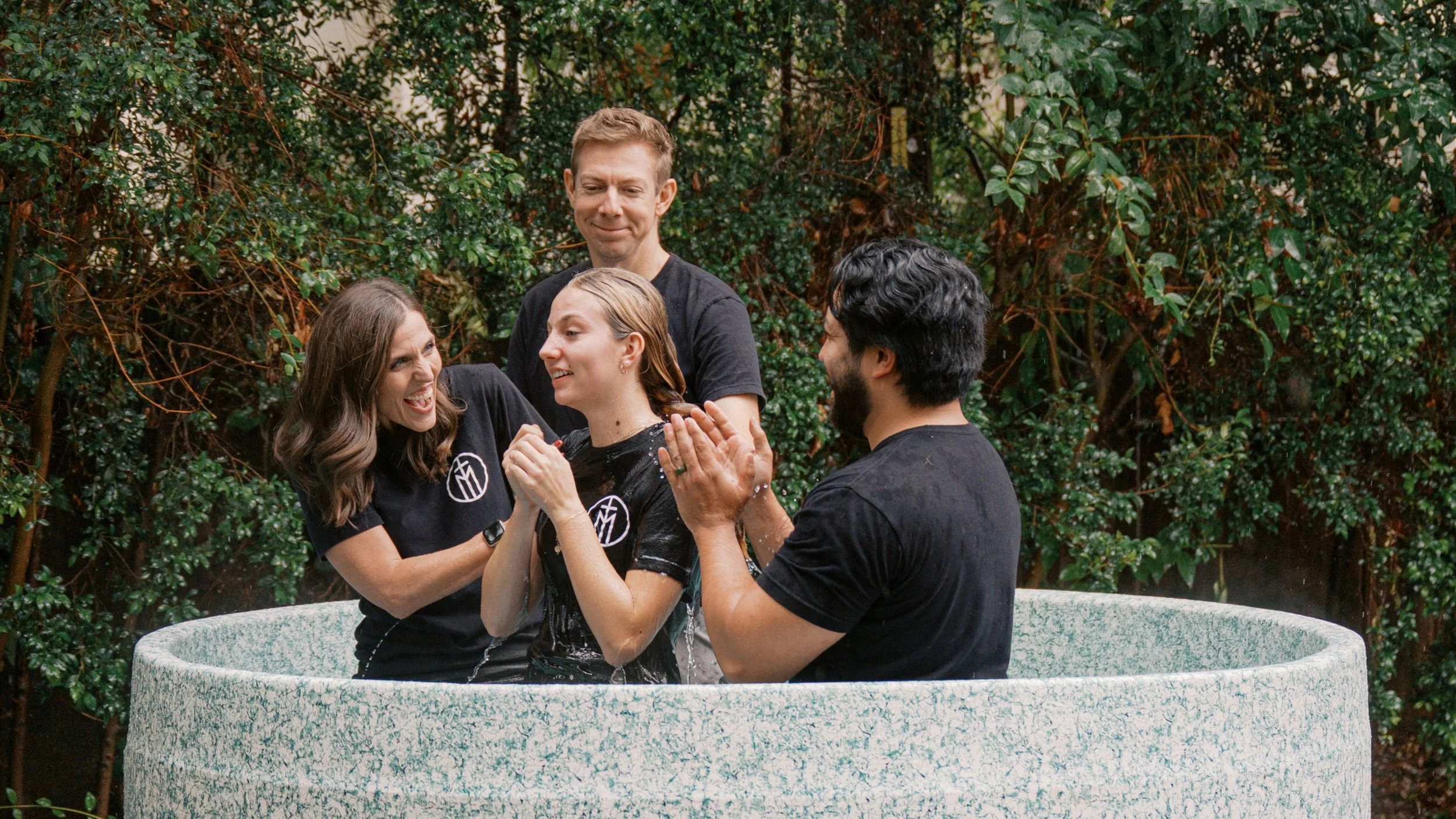 Four people having a water baptism in an outdoor, green leafy setting. Two women and two men are smiling and celebrating, with water splashing as they participate in the baptism ritual.
