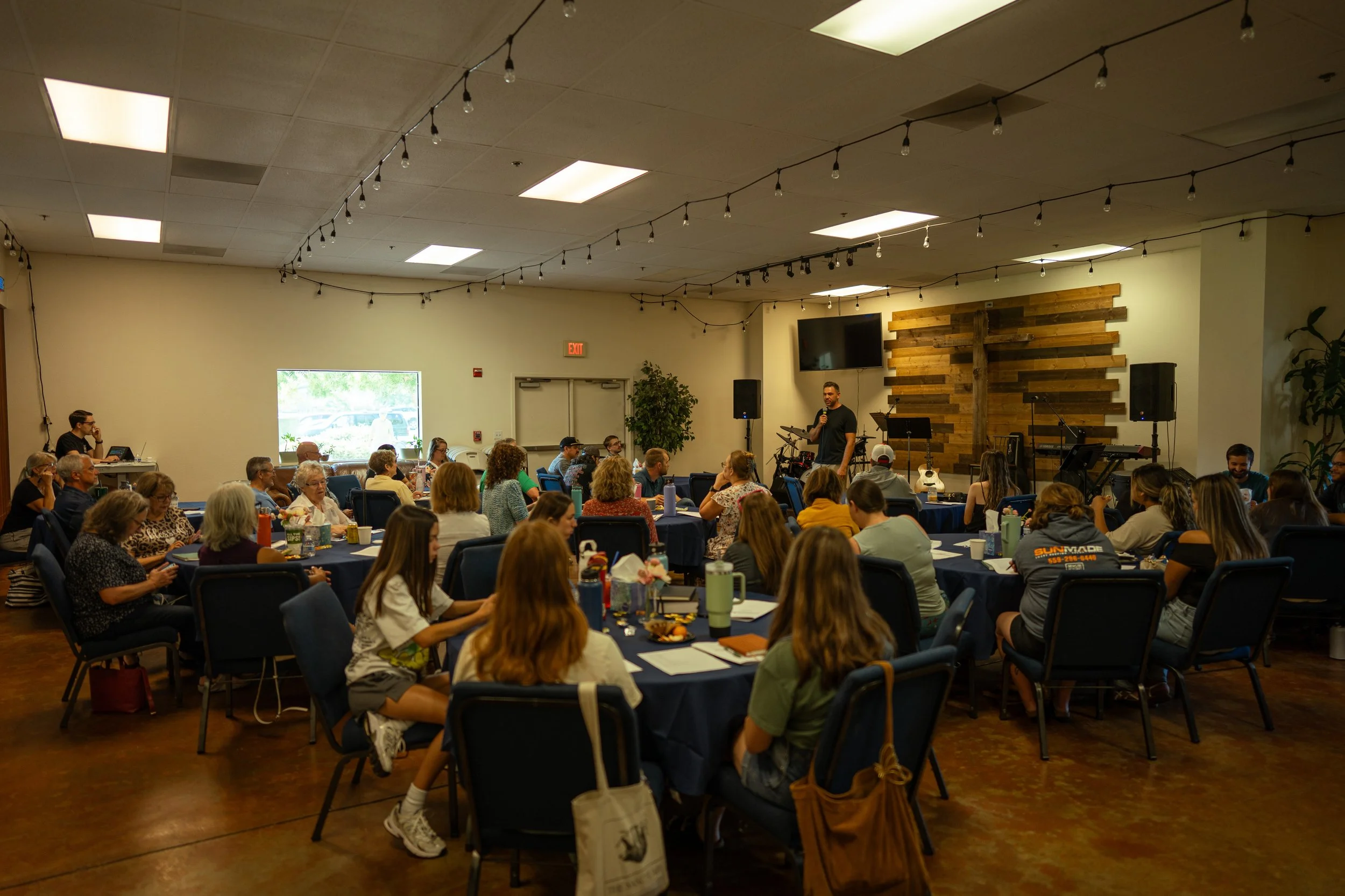 A large gathering of people sitting around tables in a community room, listening to a speaker standing near a wooden cross on the wall. The room has string lights hanging from the ceiling and a large window allowing natural light.