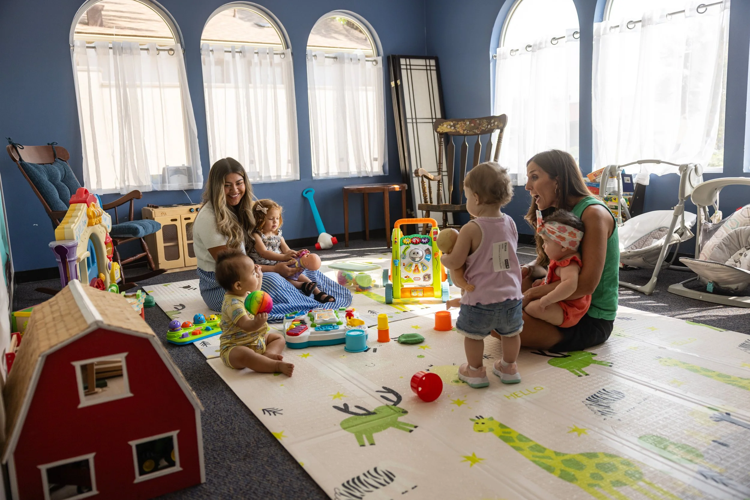 A daycare or playroom filled with young children and two adult women. The children are playing with various toys on a colorful rug, and the women are engaging with the children. The room has large windows with white curtains, and there are chairs and toys around.