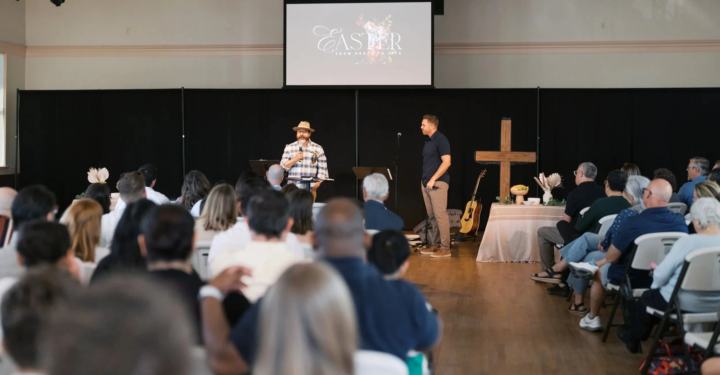 Indoor church or event space with a stage and a congregation listening to two men speaking. One man wears a hat and plaid shirt, the other wears a navy shirt. There is a large cross on the stage, a guitar, and a table with flowers.