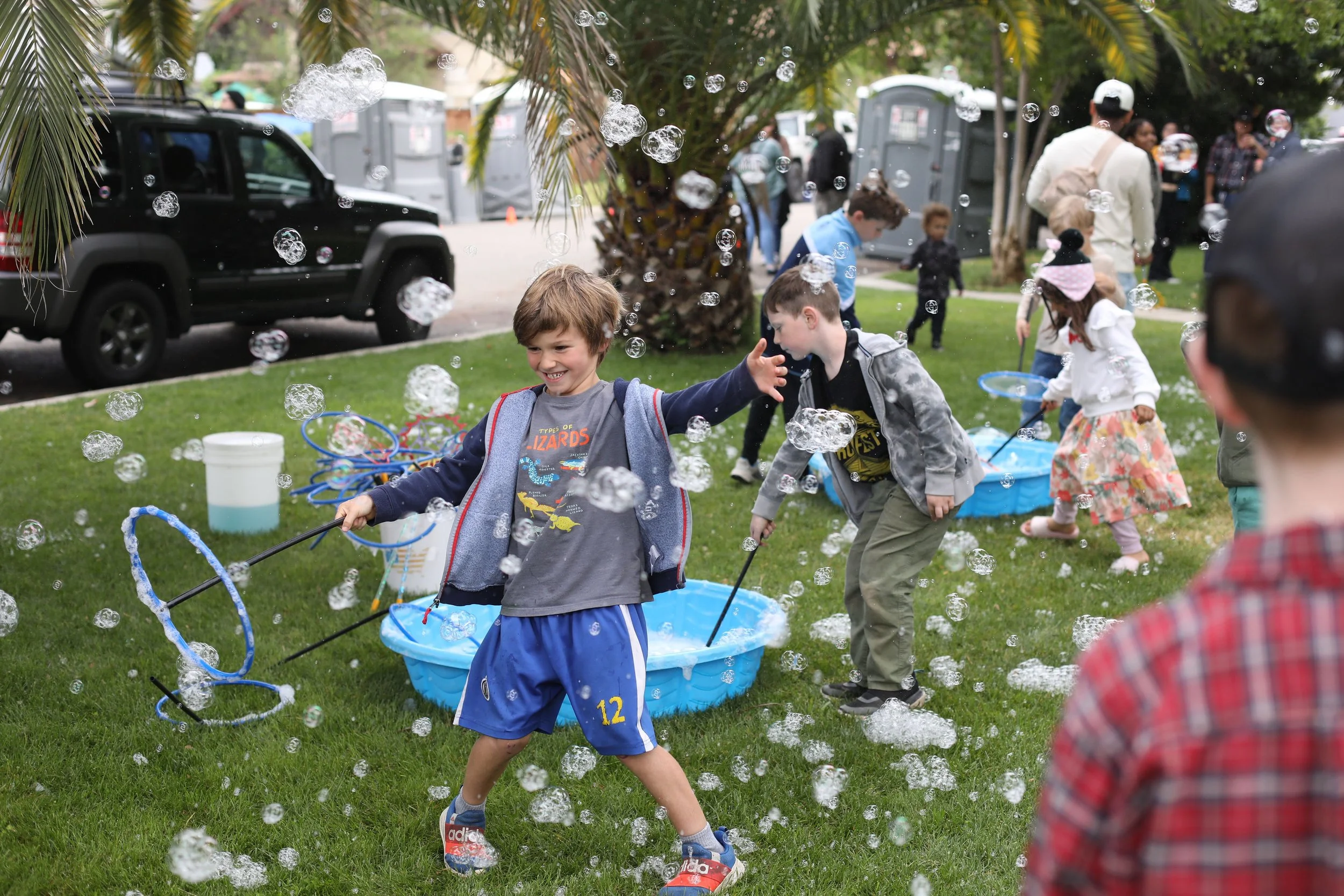 Children playing with bubbles outdoors in a park with trees and cars in the background.