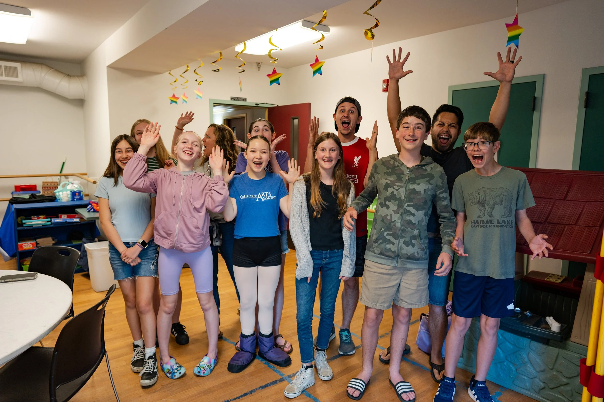 Group of children and a few adults smiling and waving at a social gathering in a room decorated with colorful rainbow star and spiral hanging decorations. Everyone appears happy and excited.
