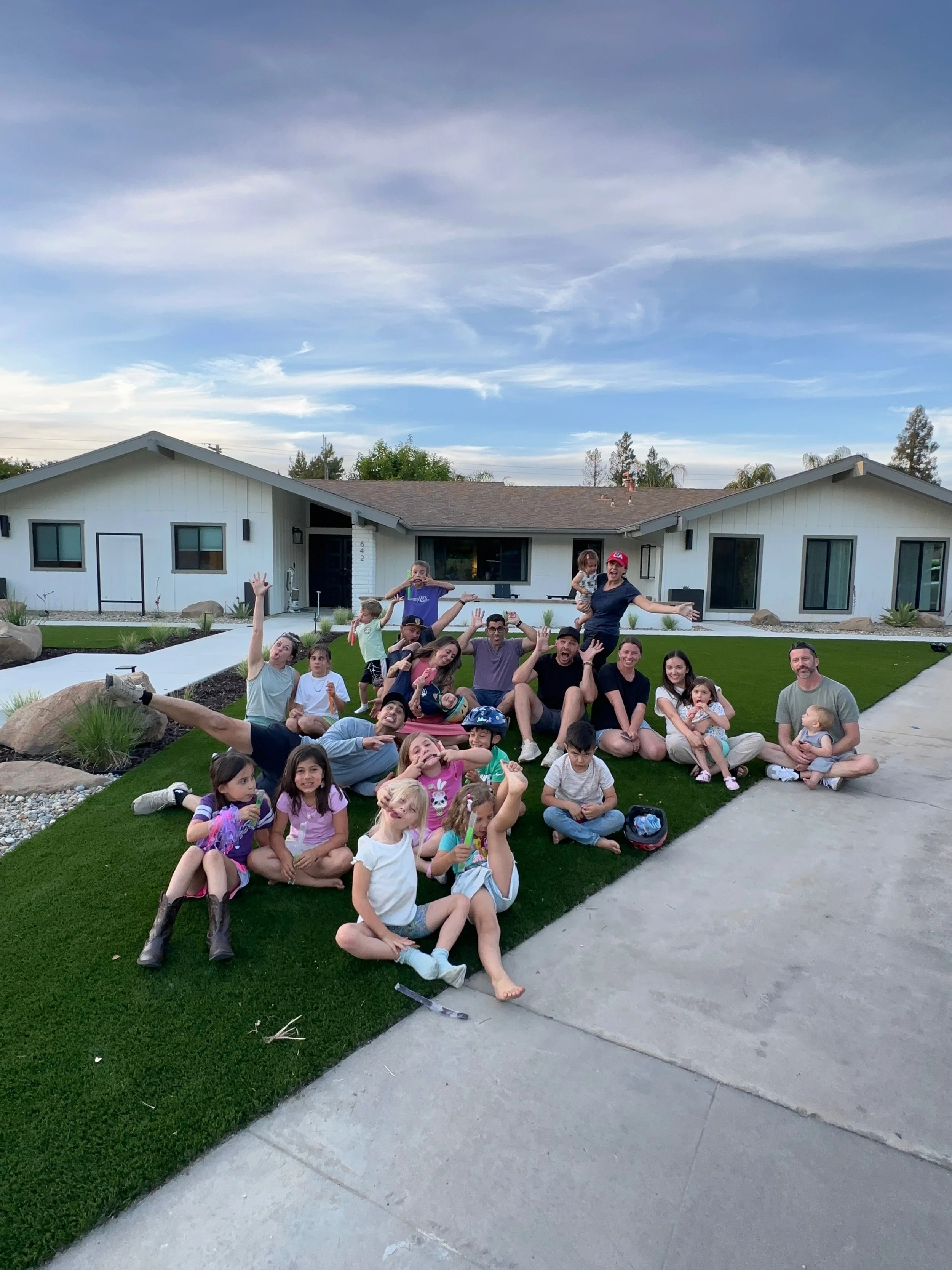 A group of children and adults sitting and lying on a well-maintained lawn in front of a modern, single-story white house with a gray roof, under a partly cloudy sky during daytime.