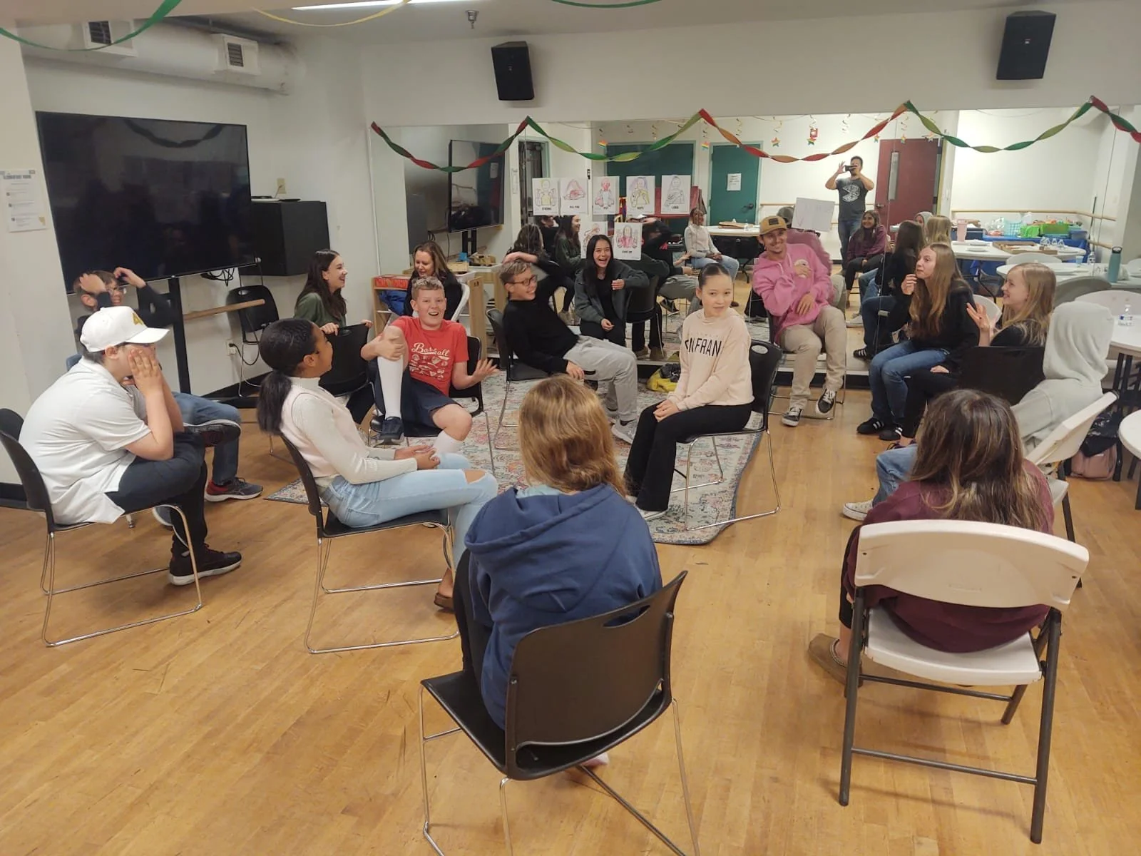 Group of people sitting in a circle in a room, laughing and engaging in a discussion or activity, with colorful decorations and drawings displayed in the background.