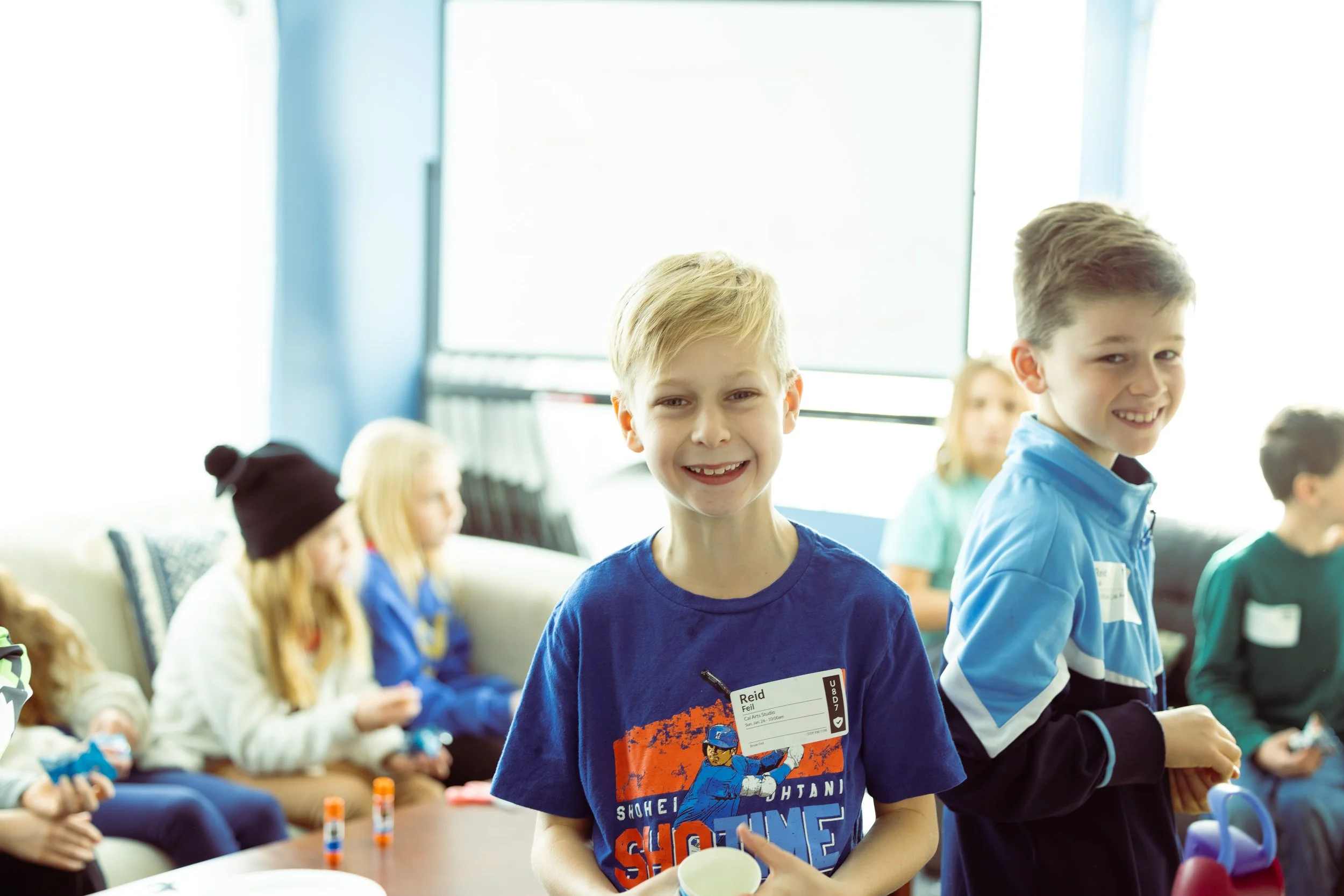 Smiling young boys in a classroom or daycare setting, standing with other children seated around a table, during a daytime activity.