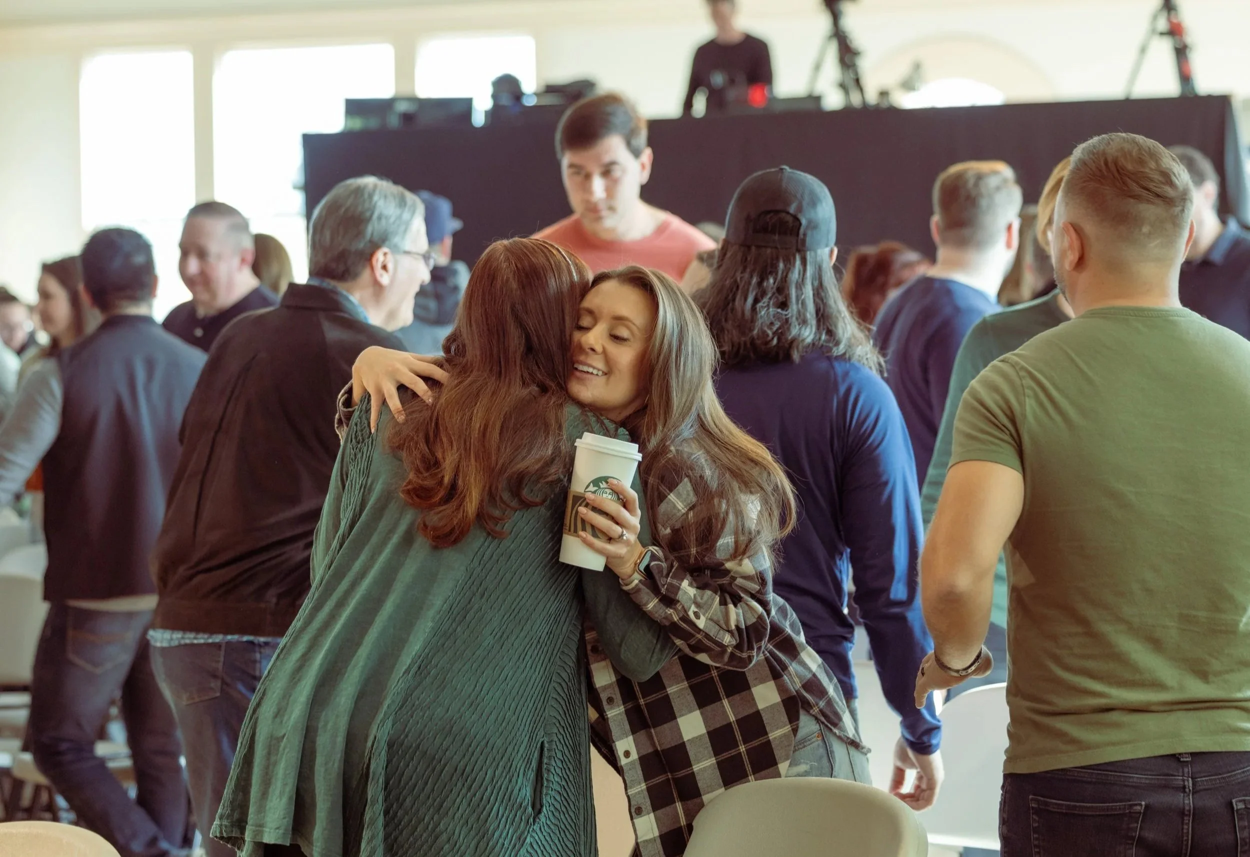 People hugging and talking at a crowded indoor event.