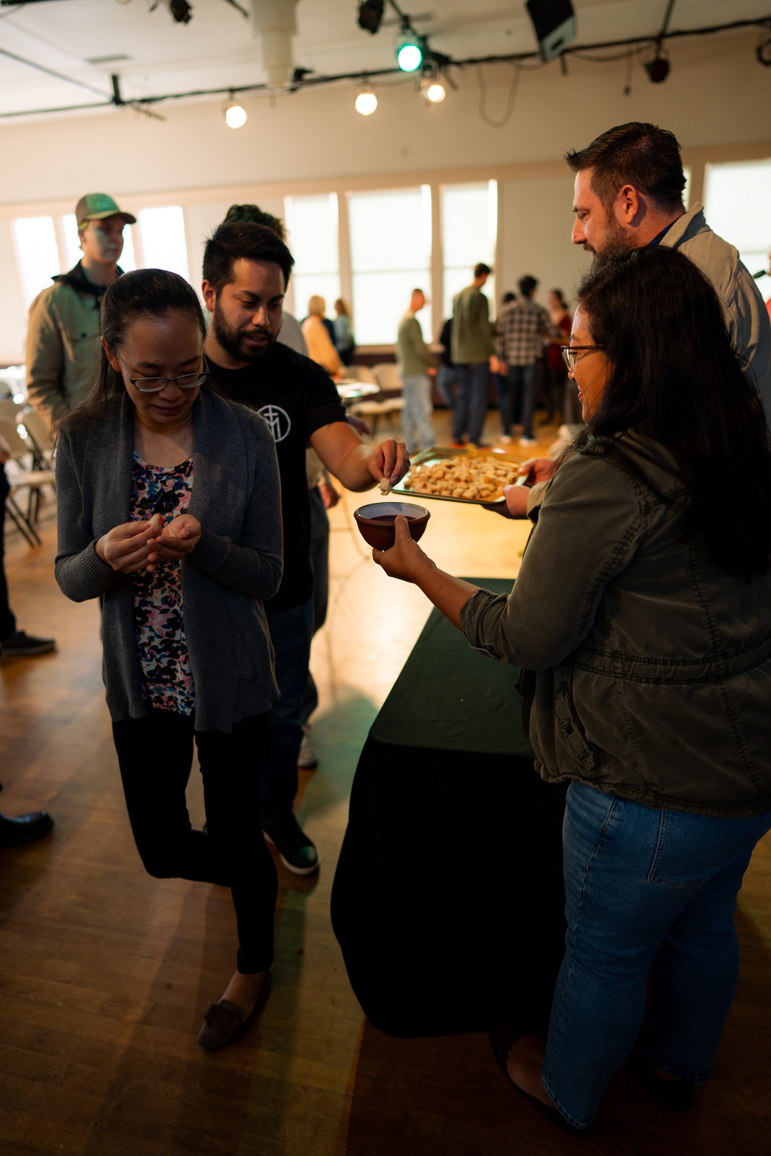 People gathered around a table at a social event, with some holding snacks and a small bowl, inside a room with large windows and a wooden floor.