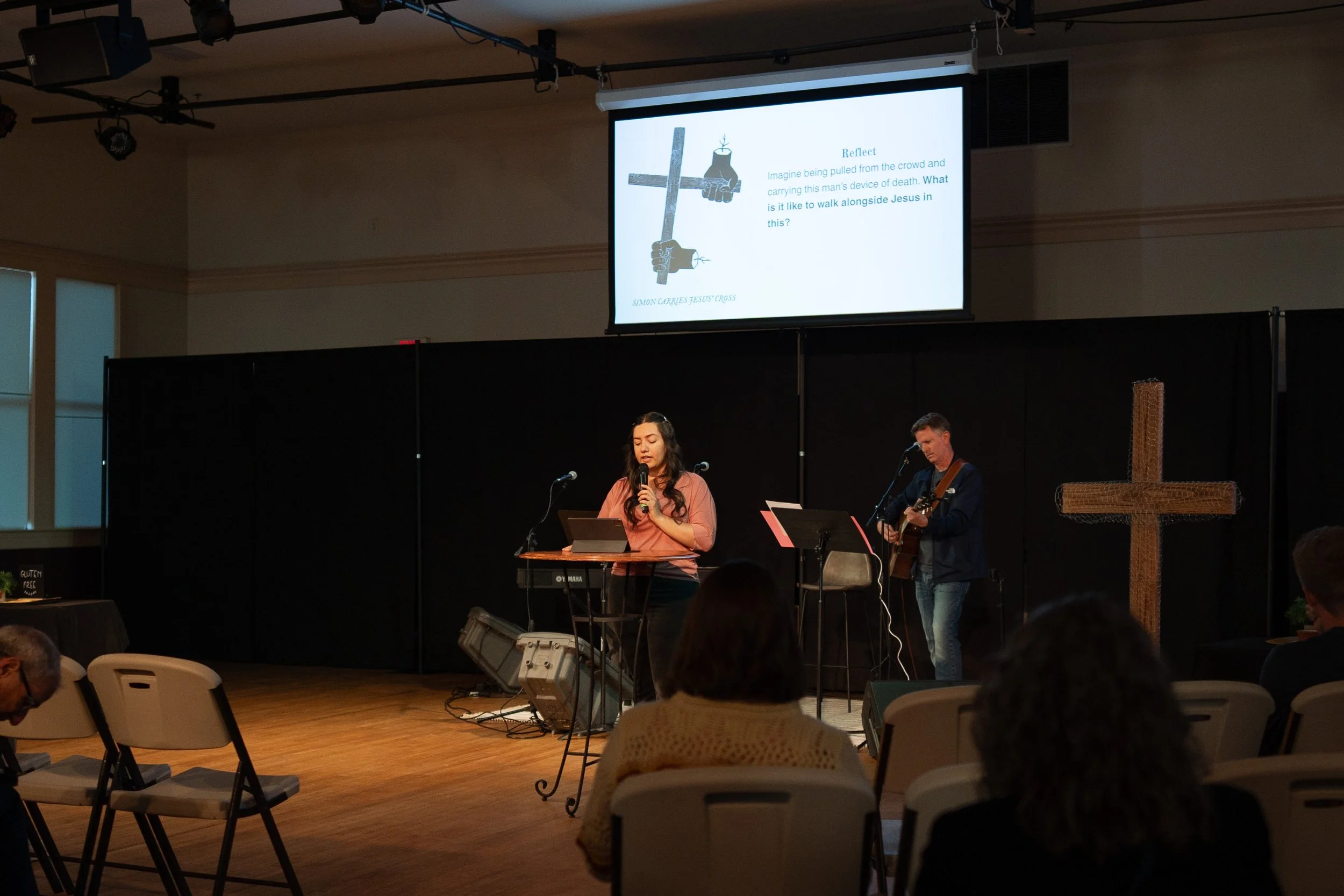 A woman singing into a microphone on stage, with a man playing guitar behind her, in a church or community hall with a cross on the right side and a large screen displaying a slide about Jesus' cross.