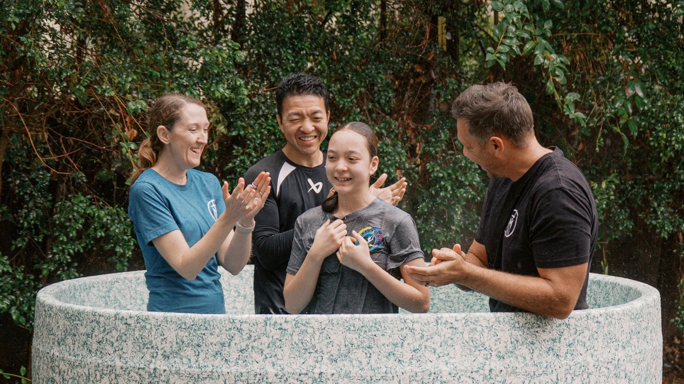 Group of five people, consisting of two men, two women, and a girl, standing in a circular stone tub outdoors, smiling and engaging with each other amidst green foliage.