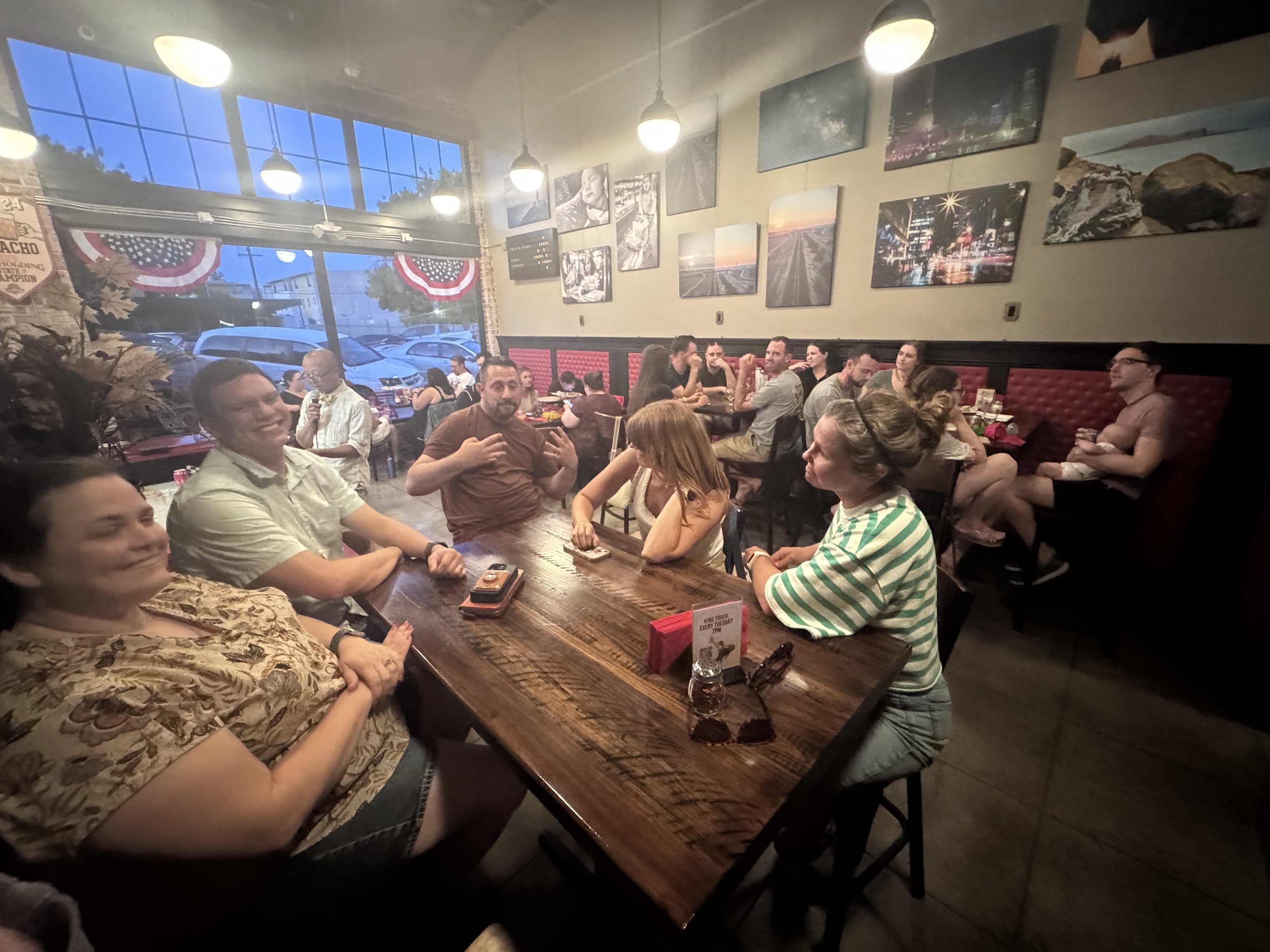 Inside a busy restaurant with patrons sitting at tables, looking at the camera, talking, and eating. Large windows with American flags and street views outside. Wall art featuring cityscapes and landscapes.