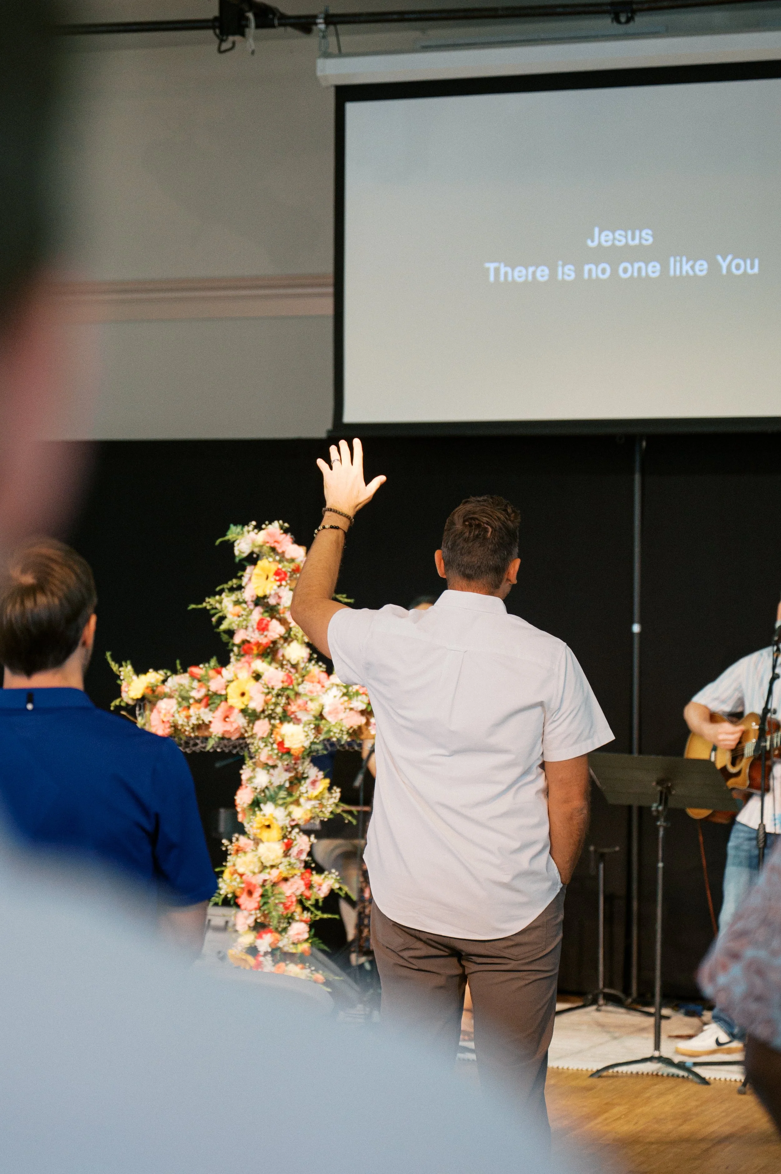 A man with a short hairstyle and wearing a white shirt is standing with his back to the camera, raising his hand during a church service. The screen displays a song lyric, "Jesus There is no one like You." There are other people in the background, and a guitarist is visible to the right. A large floral arrangement is near the front of the stage.
