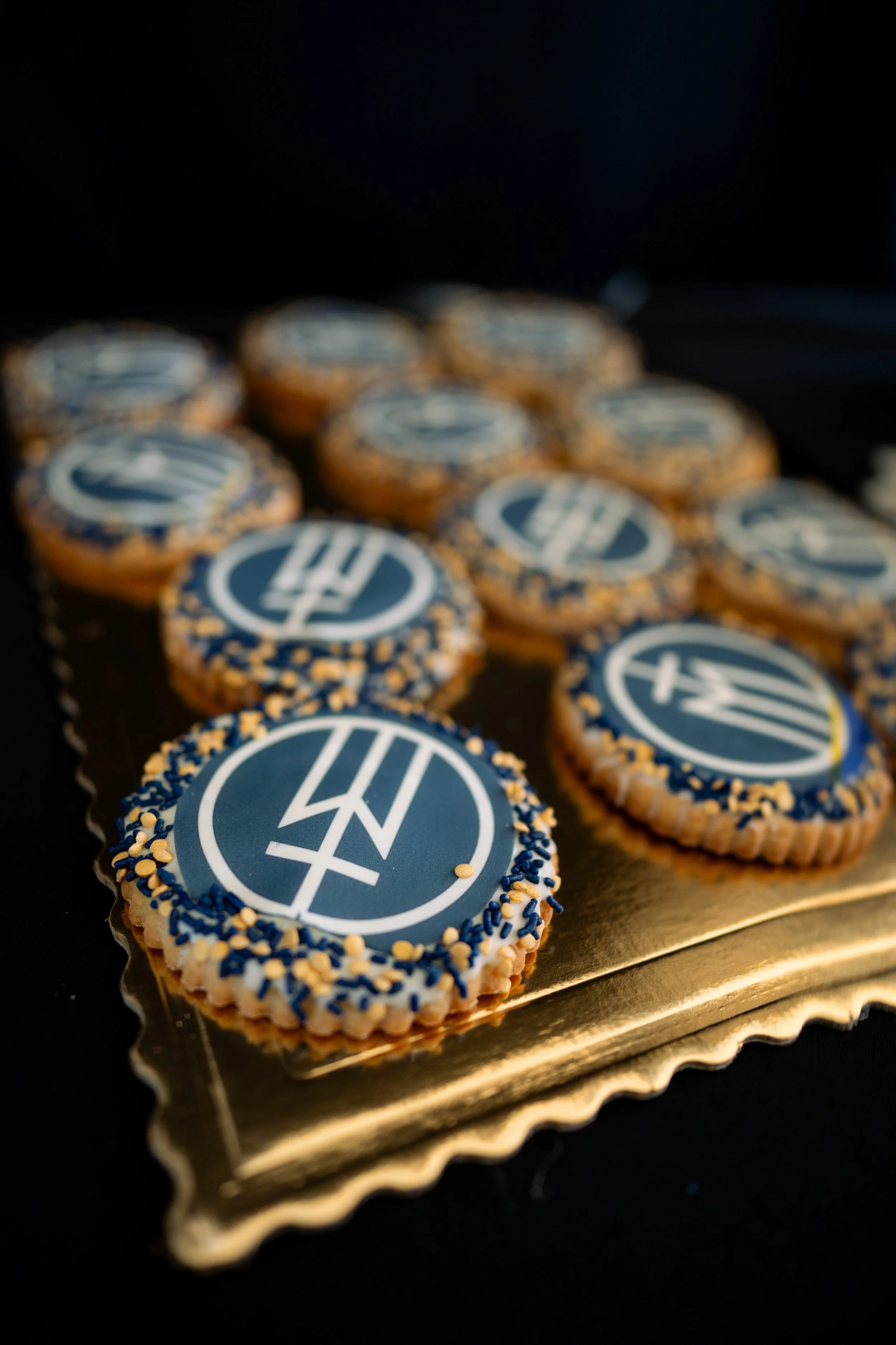 Decorated cookies with a blue and white emblem, arranged on a gold tray on a black background.