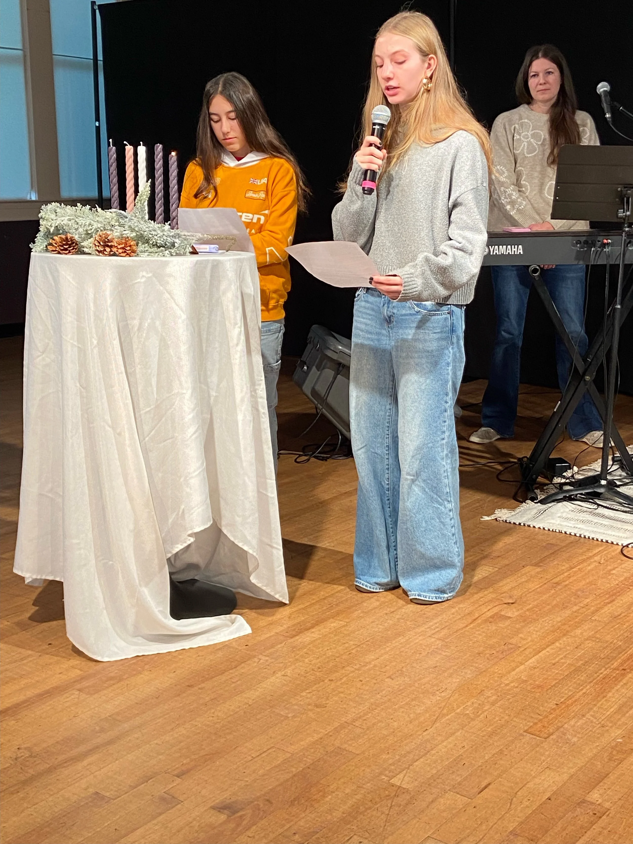 Three women standing on a wooden stage, one at a table decorated with candles, pinecones, and frosted greenery, while the other two hold papers, with a keyboard and microphone in the background.