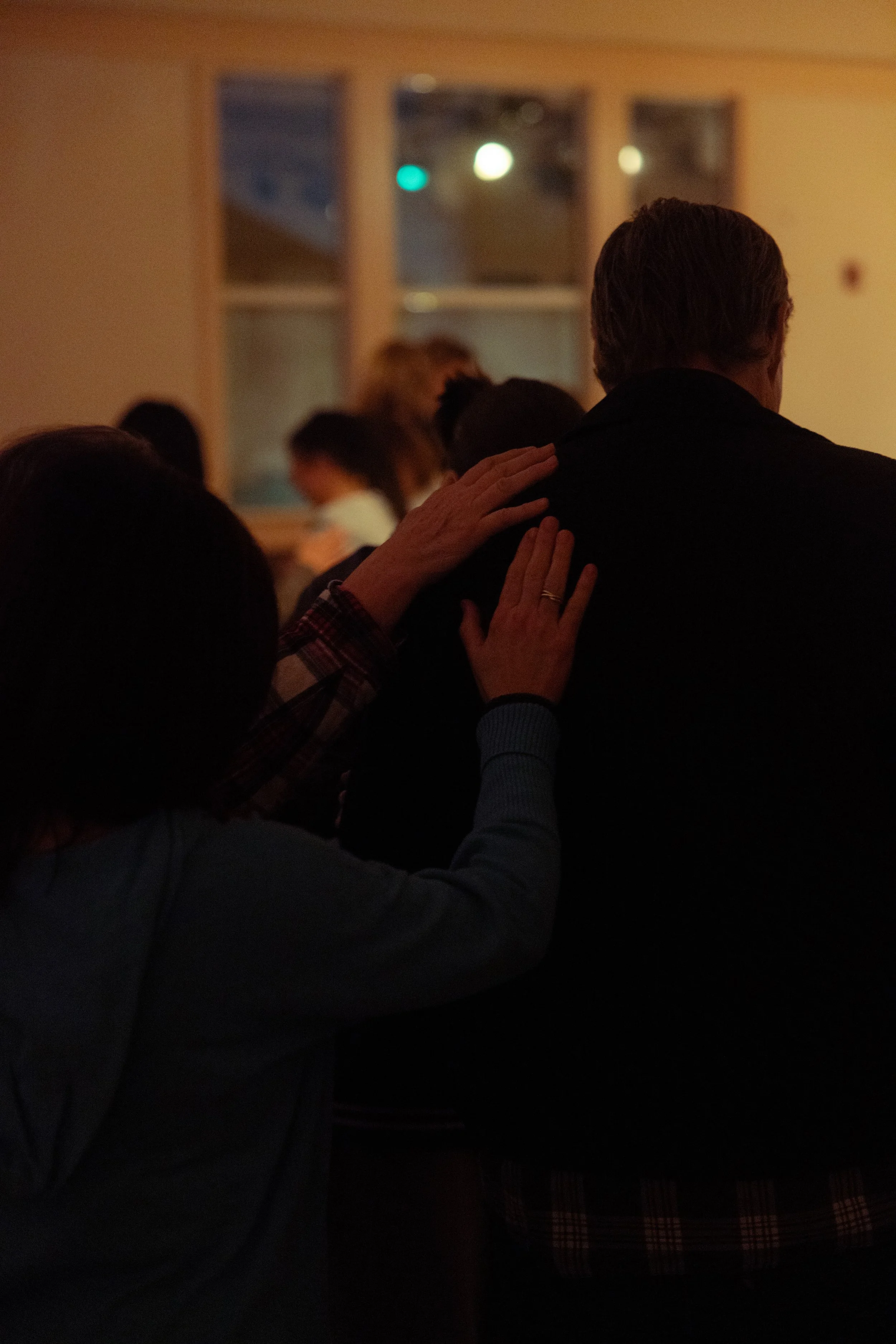 People standing close together at an indoor event, with one person placing a hand on another's shoulder, warm lighting, and blurred background.