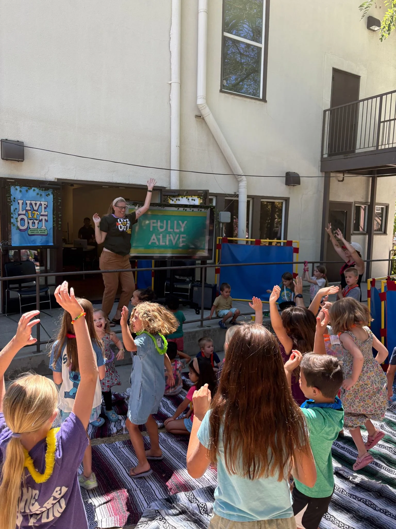 A group of children gathered outdoors, raising their hands and sitting on a striped blanket. Two women are standing at the front, leading the activity, with one waving and the other raising her hand. There is a large green banner with the words 'FULLY ALIVE' behind them, and a building with windows and outdoor lighting in the background.