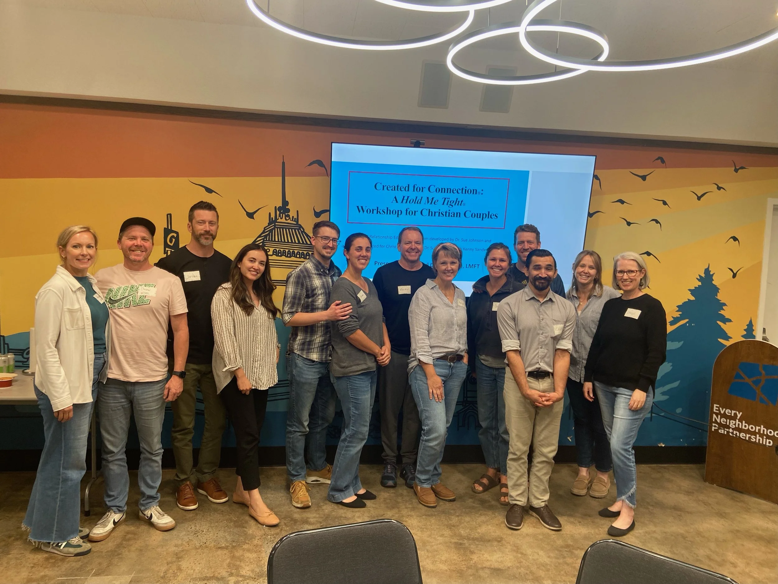 Group of twenty people, both men and women, standing together indoors in front of a screen displaying text for a workshop titled 'Created for Connection: A Hold Me Tight Workshop for Christian Couples.' The background features black bird silhouettes and a stylized cityscape. Everyone is smiling, and some people are wearing name tags.