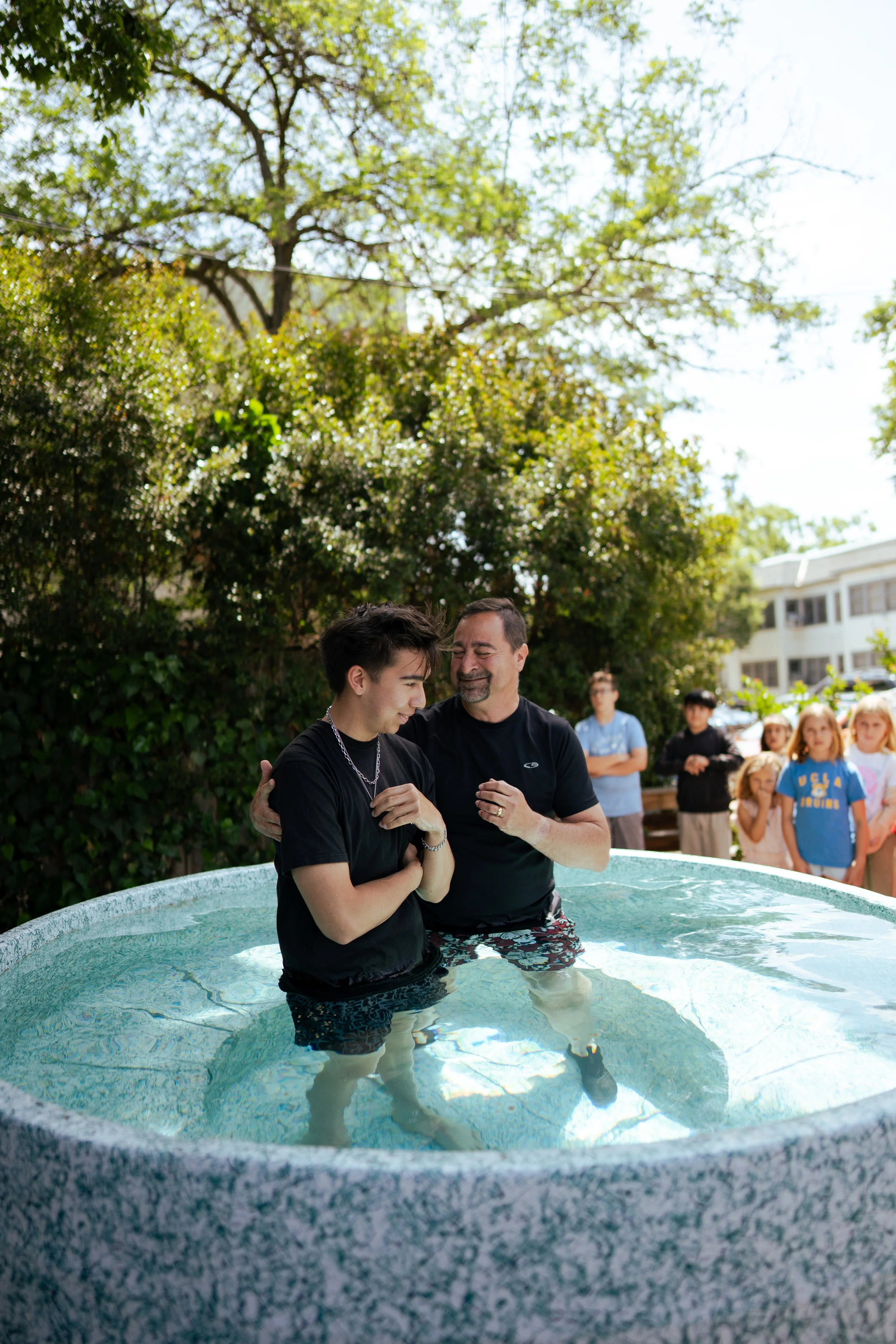 Two men standing in a small round pool outdoors, with a group of people watching in the background. Trees and a building are also visible.