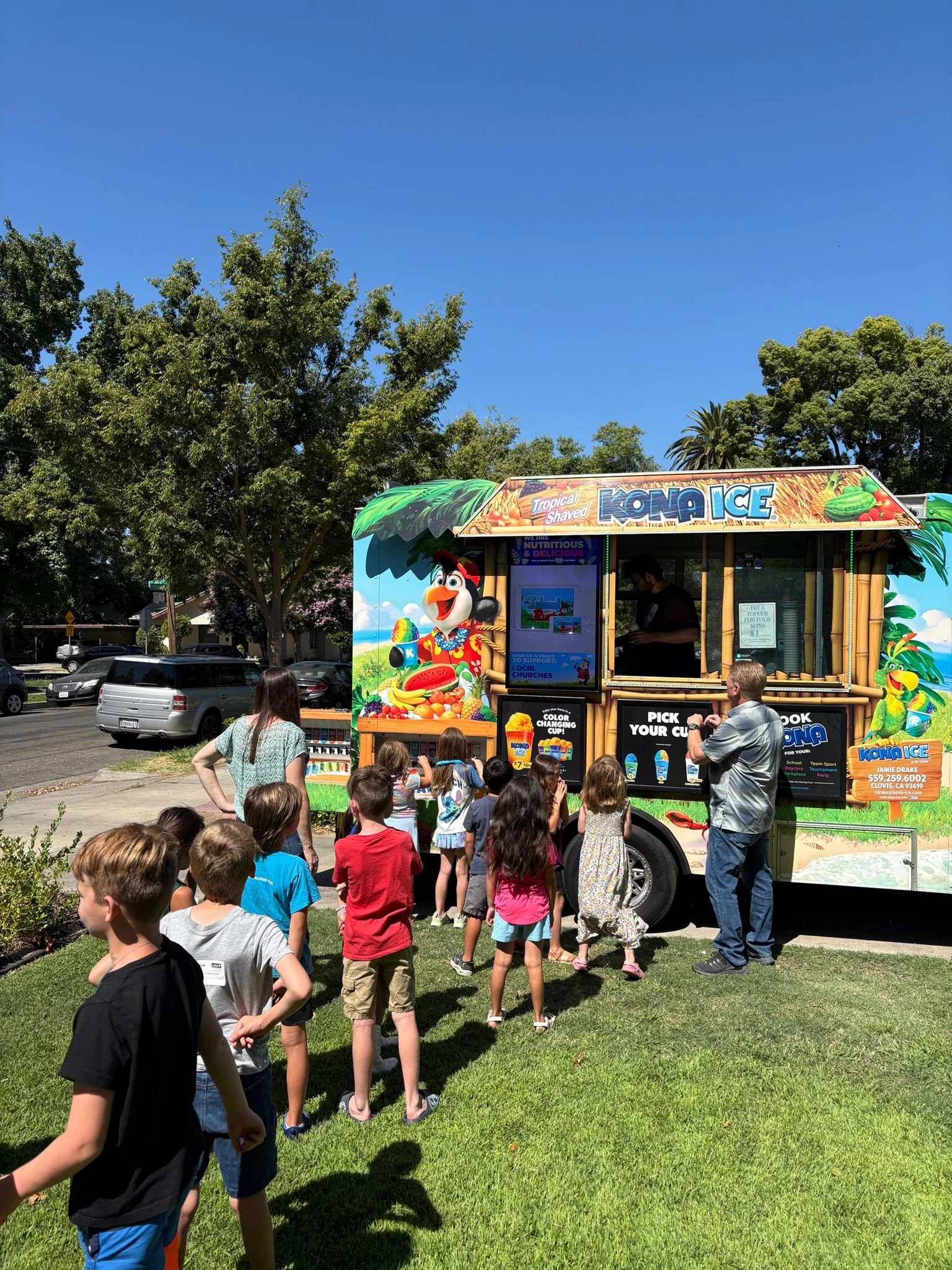 A group of children lined up outside at a Kona Ice snow cone truck on a sunny day with clear blue skies, trees, and parked cars in the background.