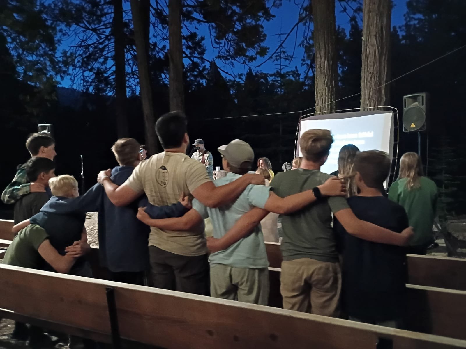 A group of people, including children and adults, standing in a semi-circle outdoors at night, watching a presentation projected on a screen surrounded by trees.