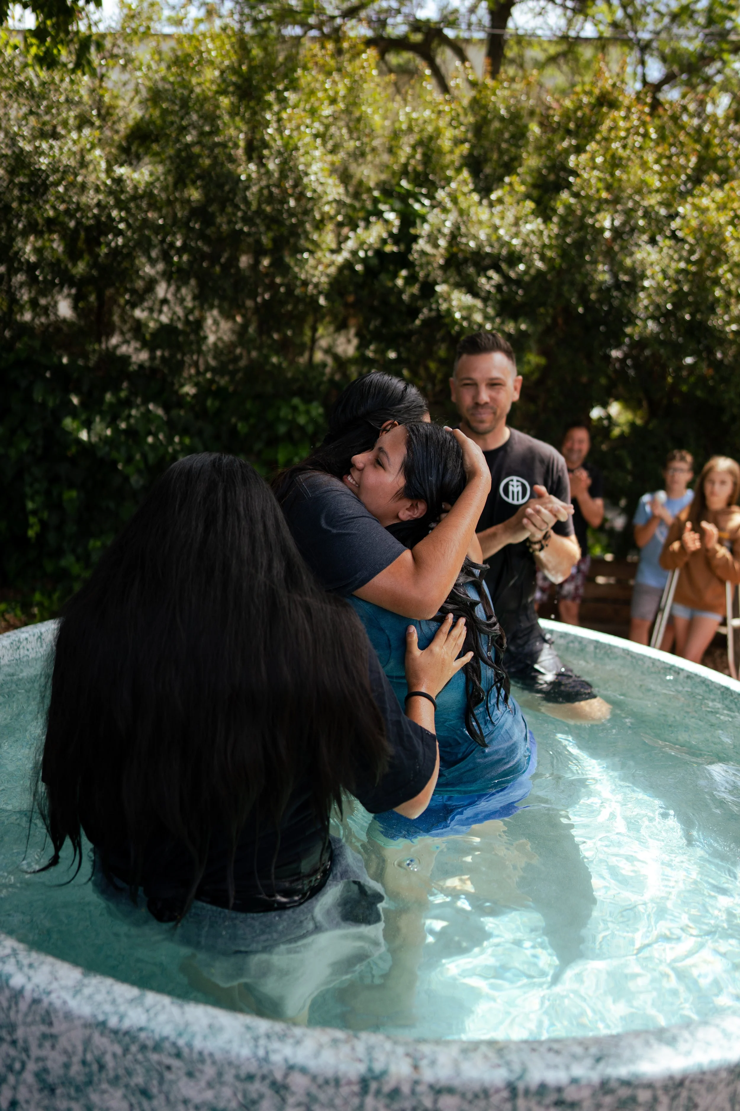 People are standing in a baptismal pool outdoors, with one person being baptized by a woman. Others are watching and clapping in the background, surrounded by trees.