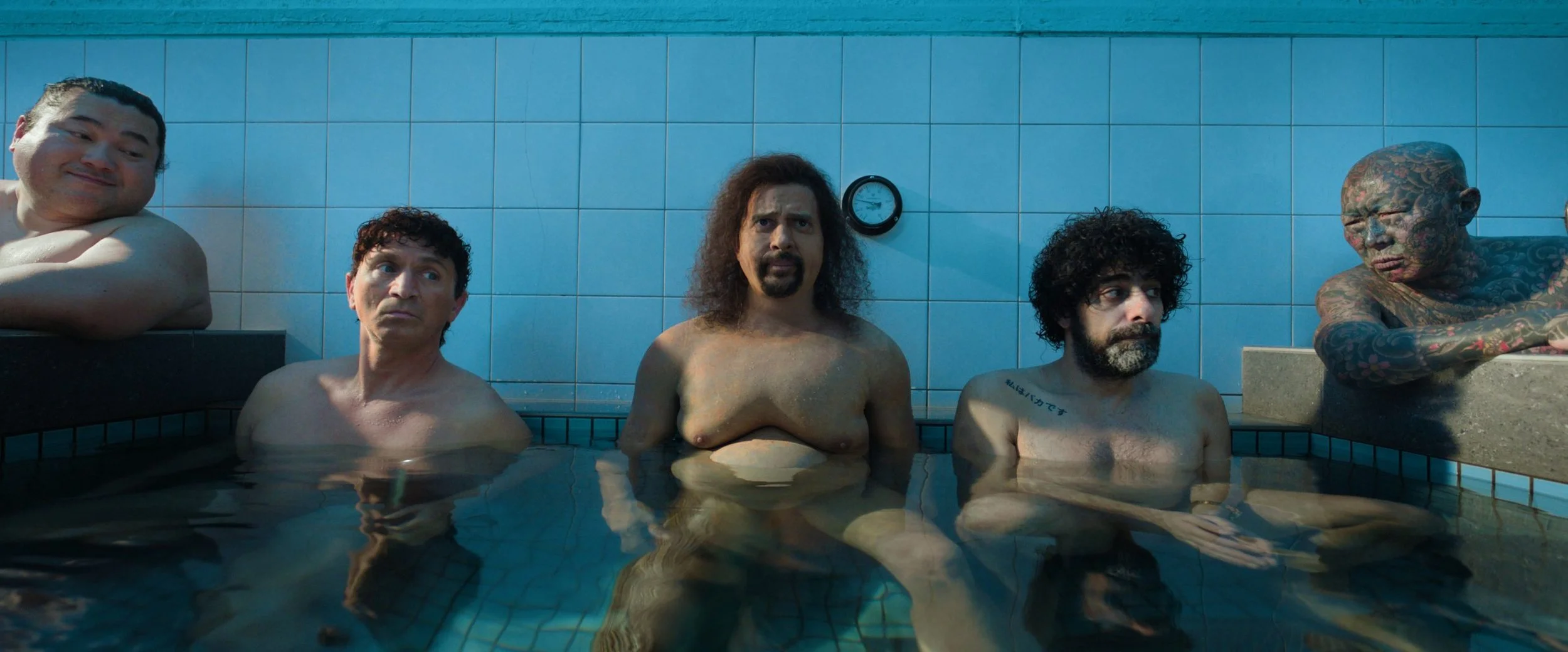 Five men in a tiled indoor swimming pool, some with tattoos and facial hair, some with curly hair, one leaning on a ledge, and one with arms crossed, all partially submerged in water.