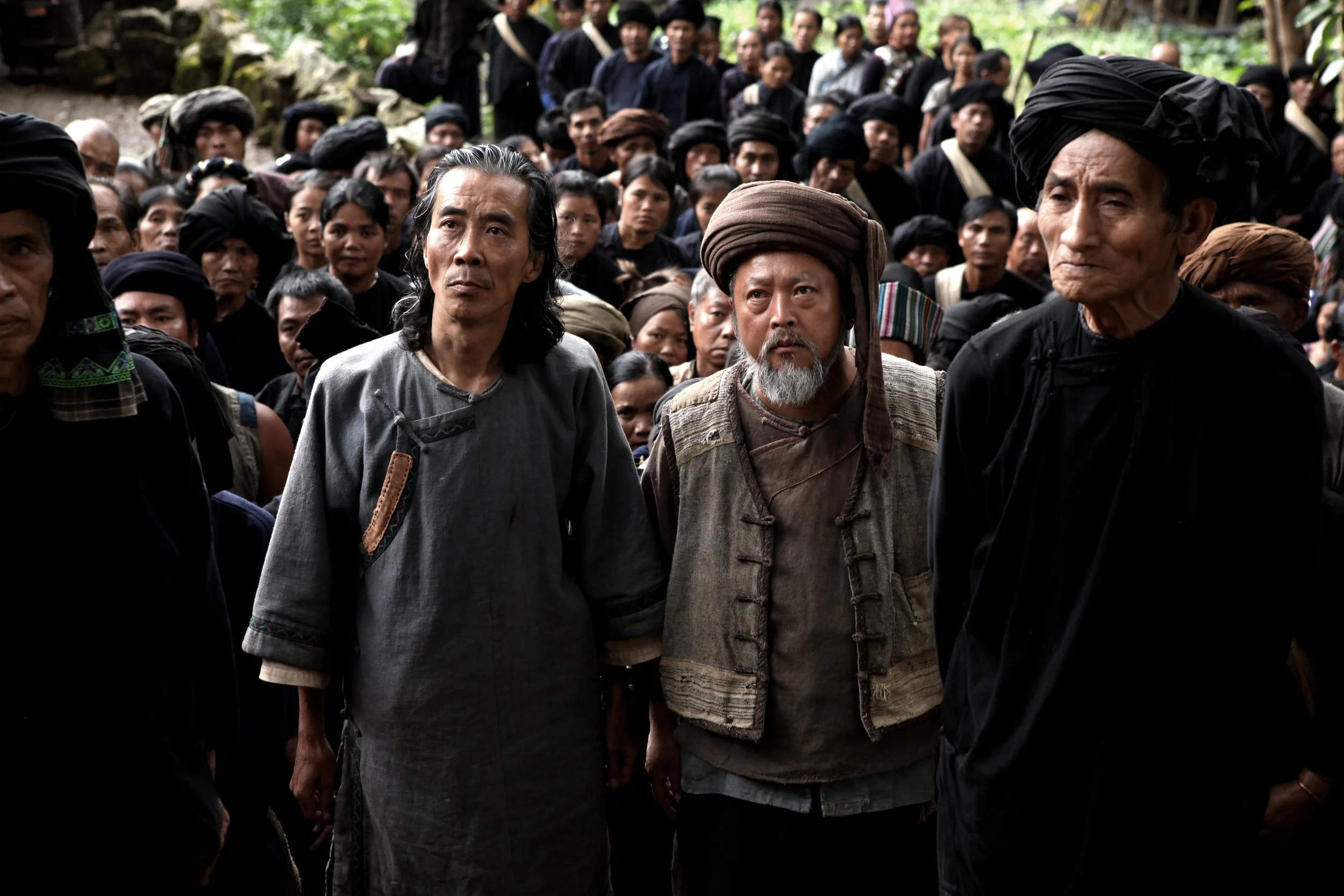 A large group of men and women dressed in traditional black clothing, gathered outdoors in a tropical setting with green foliage in the background. Some men are wearing headwraps or turbans.