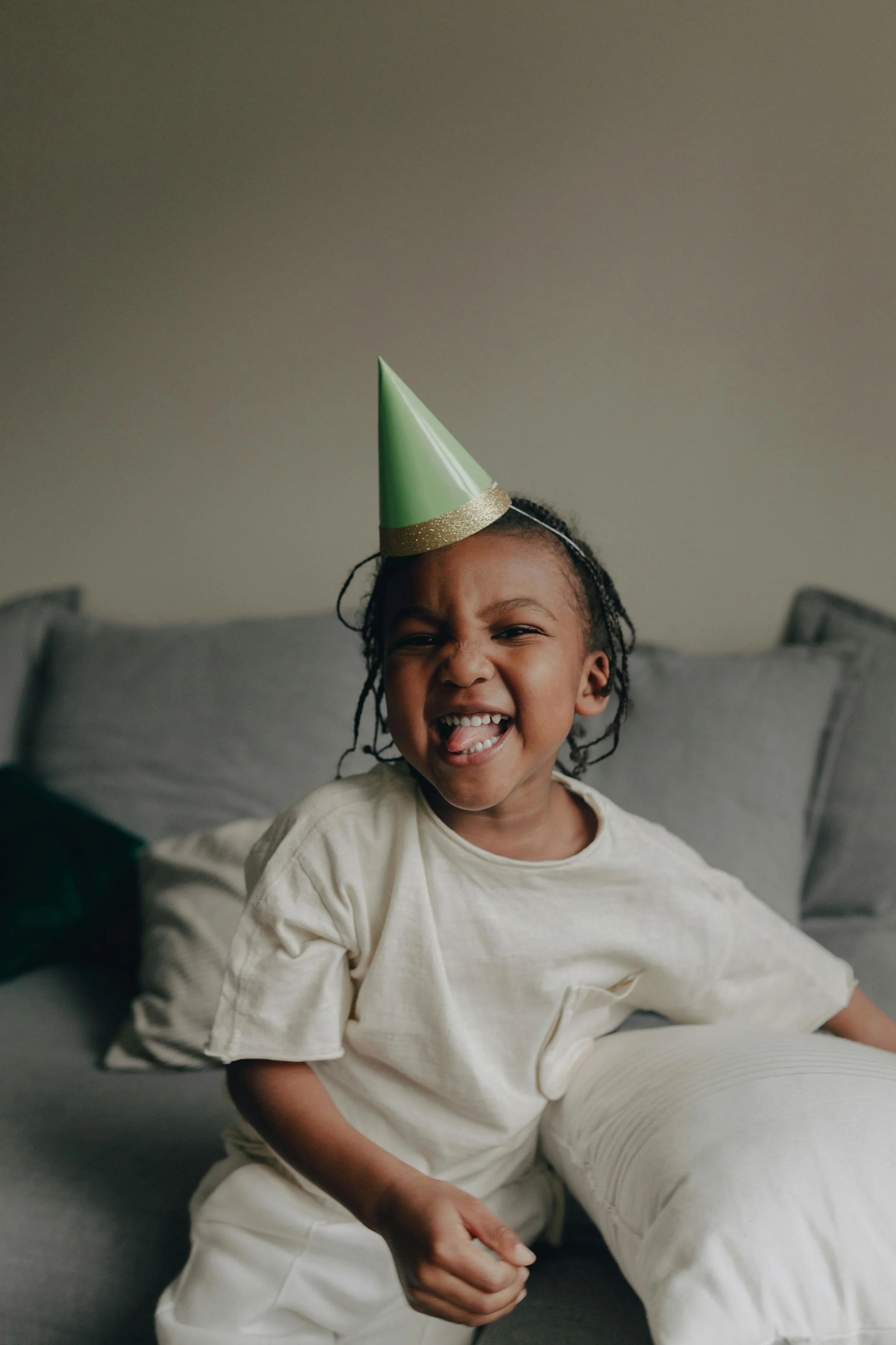 Happy child wearing a green party hat and light-colored T-shirt, sitting on a gray couch and smiling.