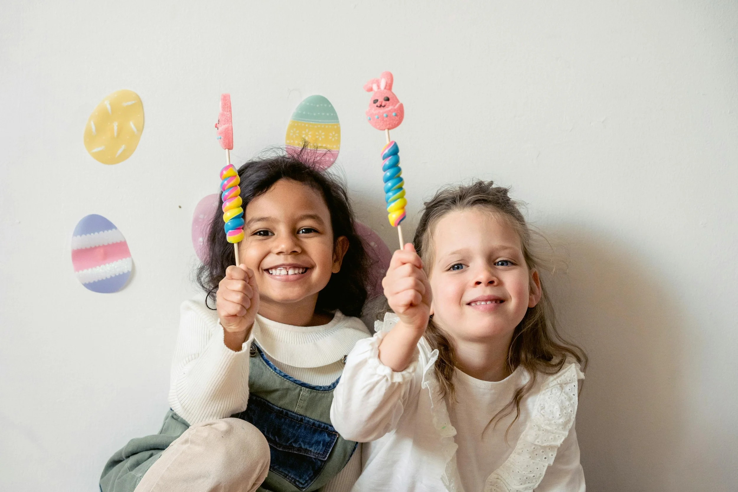 Two children smiling and holding colorful spiral lollipops with Easter-themed decorations in the background.