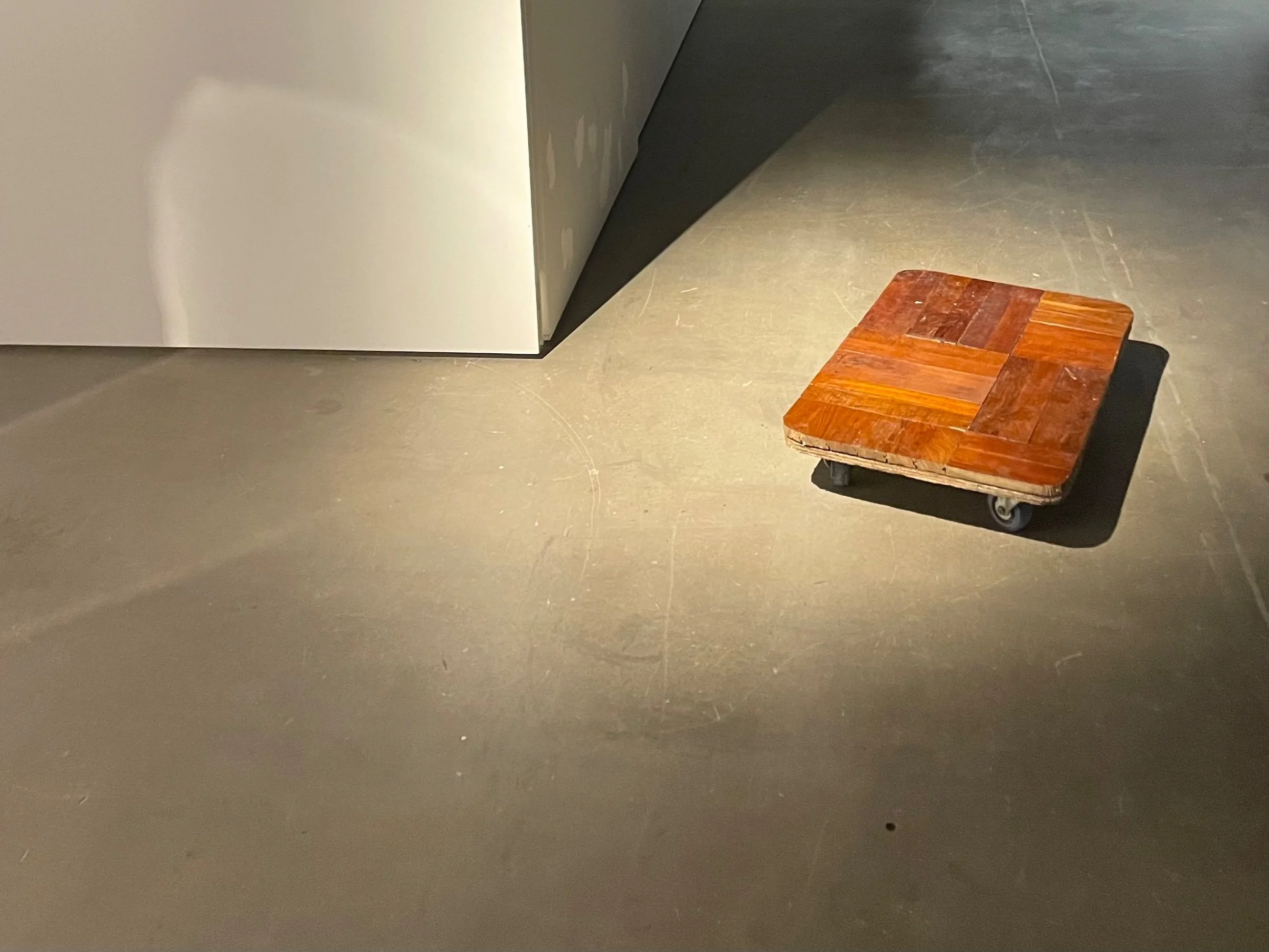 This is the work of Aidan Ng Ho Long, called 'Positioning'. A small wheeled cart or dolly was covered with teak wood flooring tiles, on a grayish concrete floor.