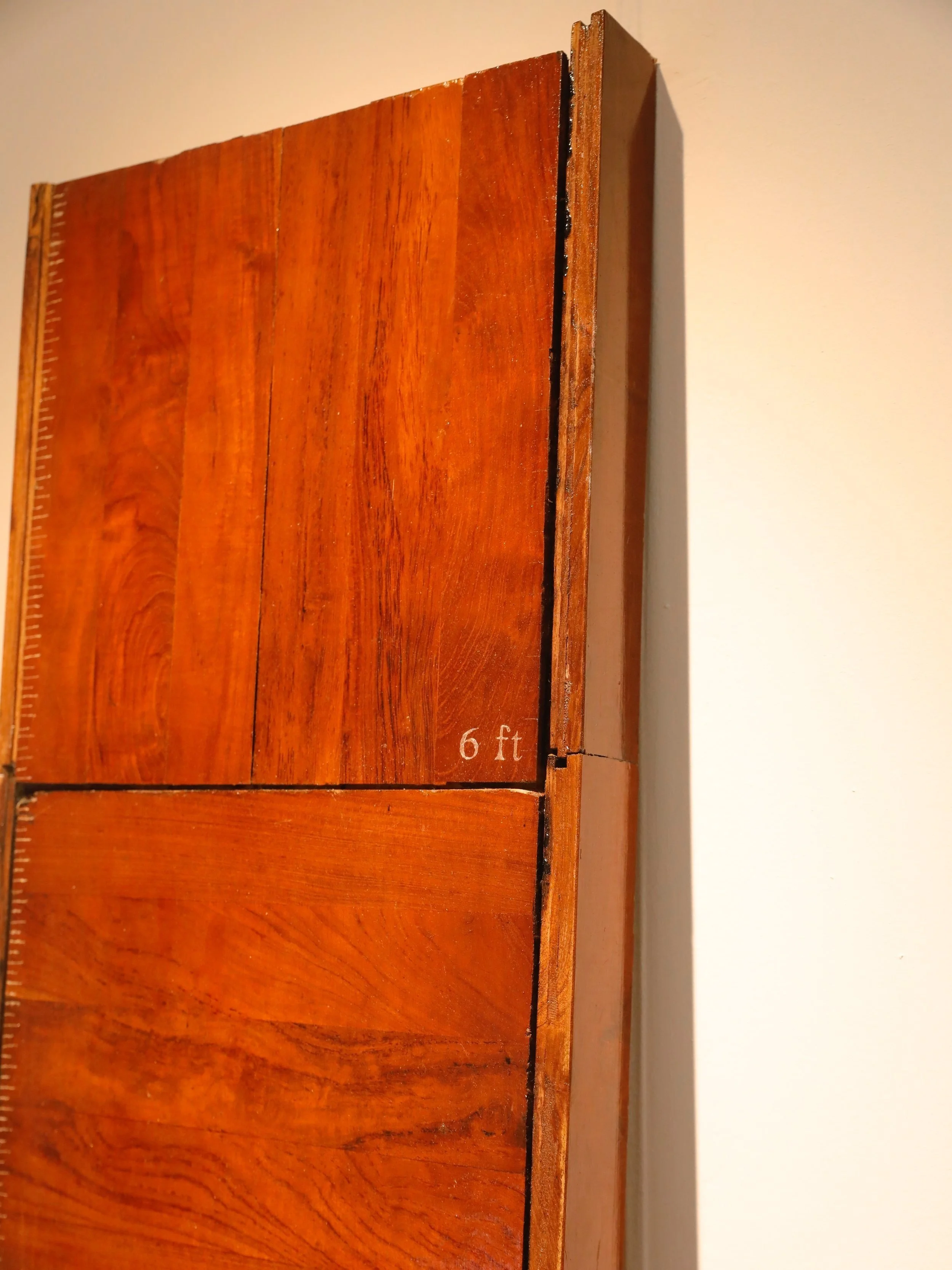 A close-up image of a wooden measuring stick on a wall, with the text '6 ft' visible near the bottom and a shadow cast on the wall.