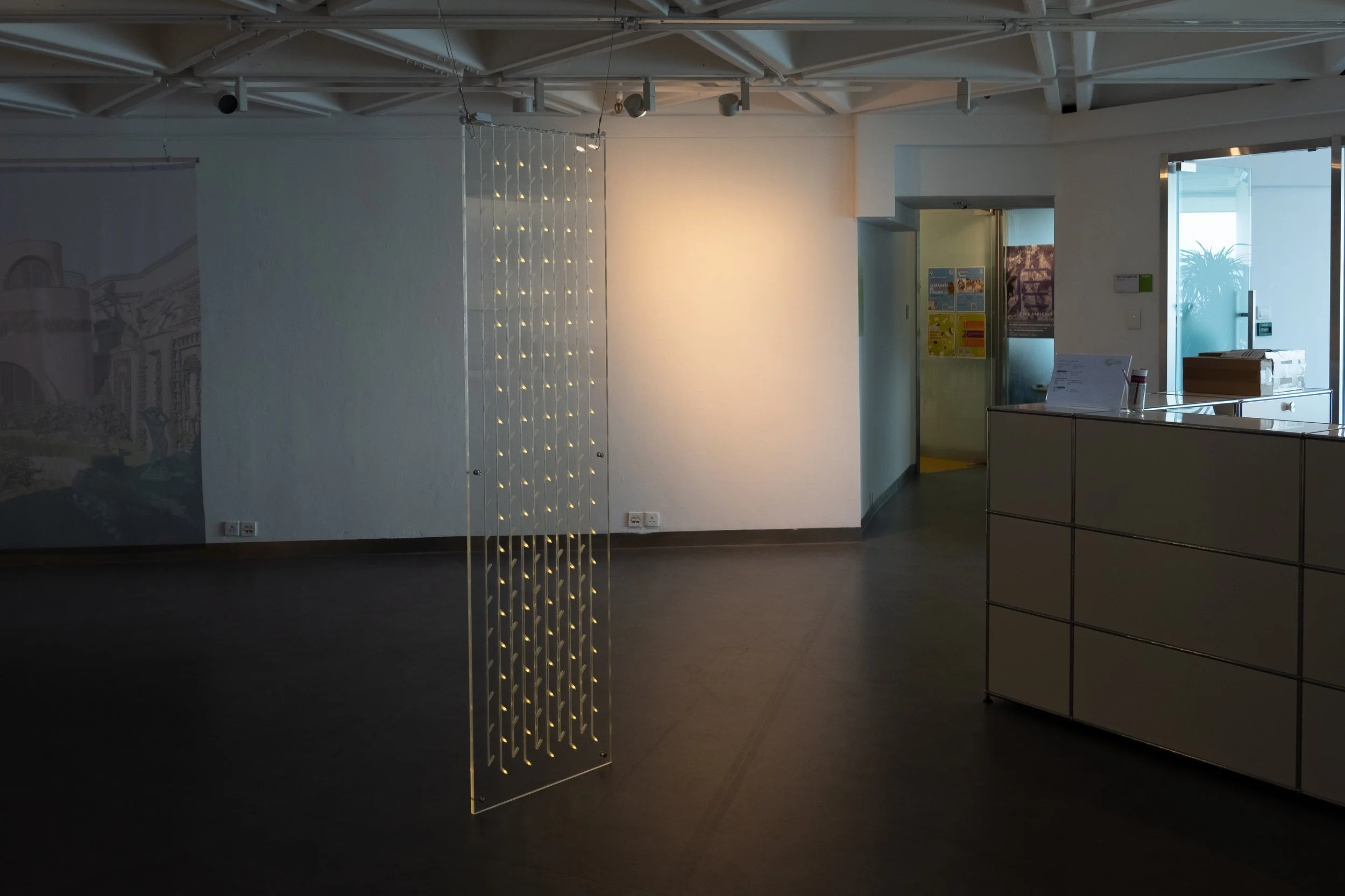 Interior of an art gallery or exhibition space with a decorative wire sculpture illuminated by small lights, white walls, and a reception desk near a glass door with posters on the wall.