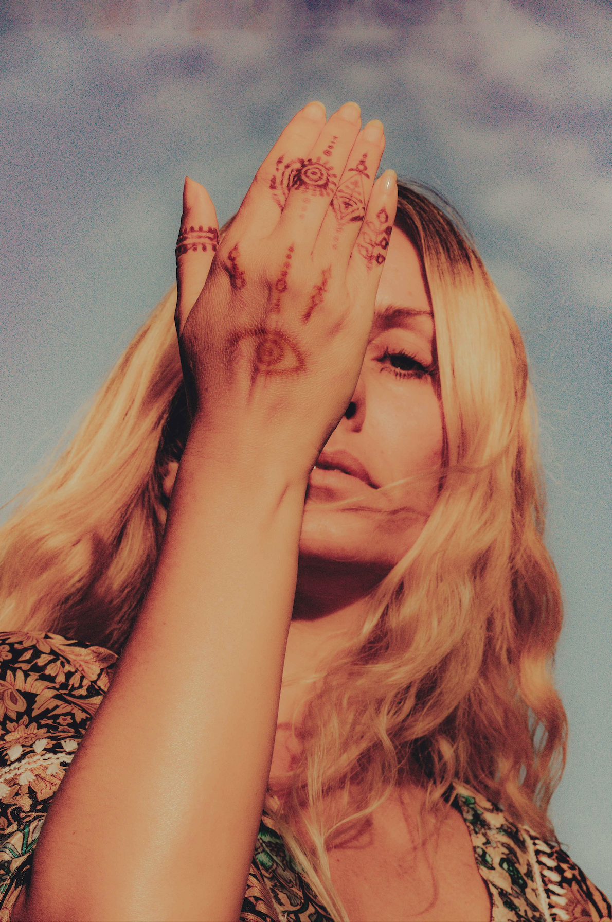 A woman with long, wavy blonde hair , marie-laure daillut, covering one eye with her hand, which has various tattoos and henna designs.
