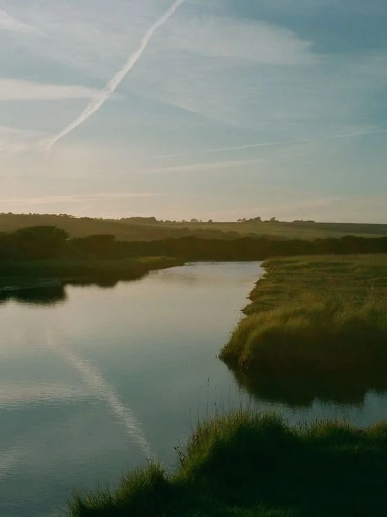 A river flowing through a grassy landscape under a sky with wispy clouds and contrails.