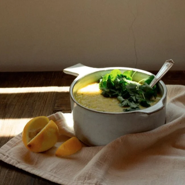 A bowl of yellow soup garnished with greens, with lemon wedges and a spoon on a beige cloth on a wooden surface.
