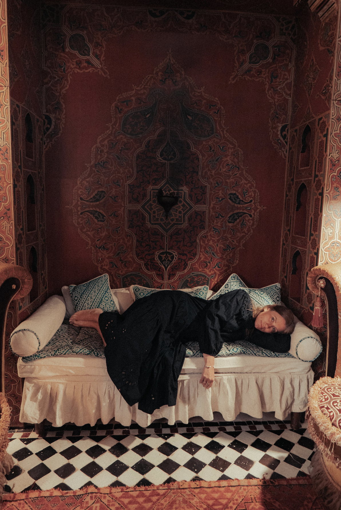 A woman lying on a patterned sofa in an ornately decorated room with intricate wallpaper and a geometric black-and-white tiled floor, wearing a black dress with her head on a pillow and looking at the camera.