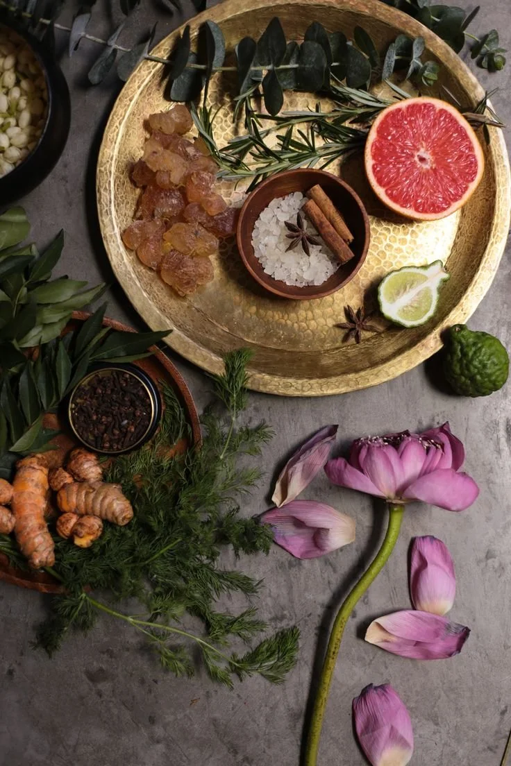 A round golden tray with a halved grapefruit, lemon wedge, pink flower petals, and herbs. Nearby are ginger roots, a small bowl of salt, a cinnamon stick, star anise, and aromatic spices, along with a small green fruit.