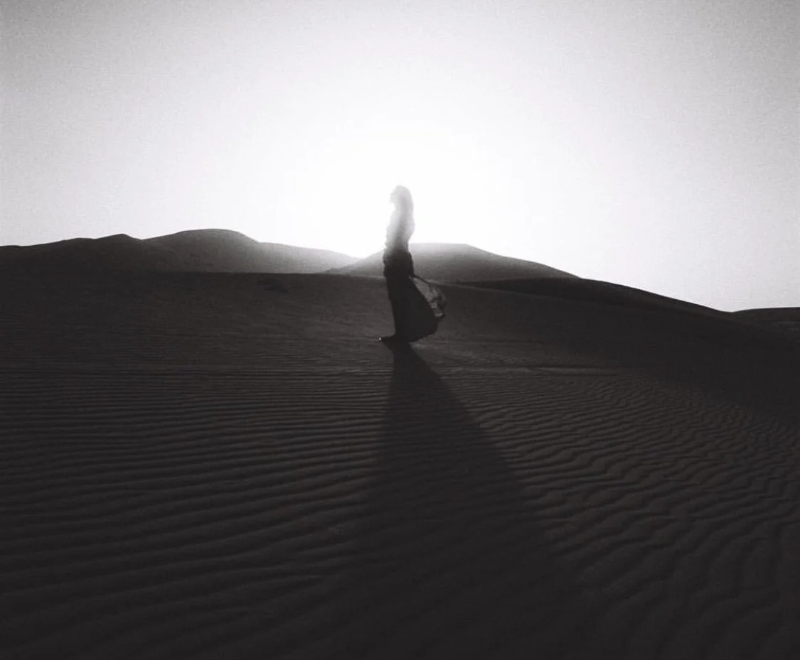 A silhouetted person walking across sand dunes at sunset or sunrise, casting a long shadow.