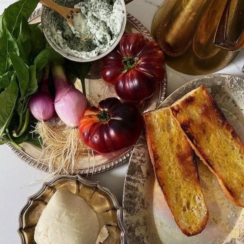 Fresh vegetables, including purple onions, green herbs, and heirloom tomatoes, along with slices of rustic bread and a dish of soft cheese on a platter.