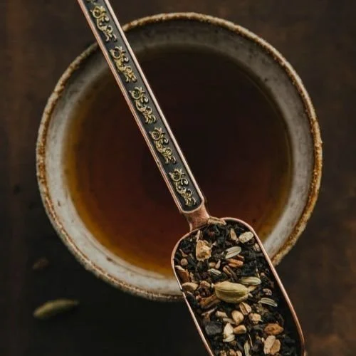 A ceramic cup filled with tea, viewed from above. There is a copper spoon resting inside the cup, containing loose tea leaves, herbs, and spices.
