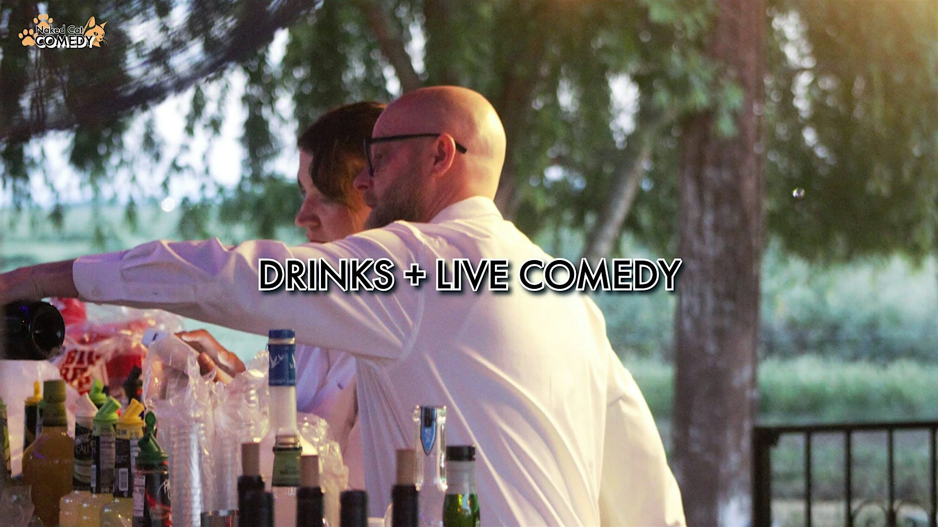 Two men at an outdoor bar, one with glasses and a bald head, the other with dark hair, surrounded by bottles and drinks, with trees in the background, under sunlight. The text overlay reads "DRINKS + LIVE COMEDY".