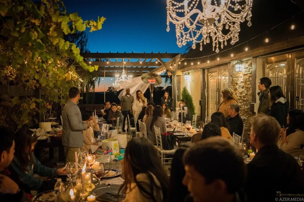 People gather for a dinner party on a decorated outdoor patio at dusk, with a large chandelier hanging above and string lights, a patio heater, and a sunset in the background.