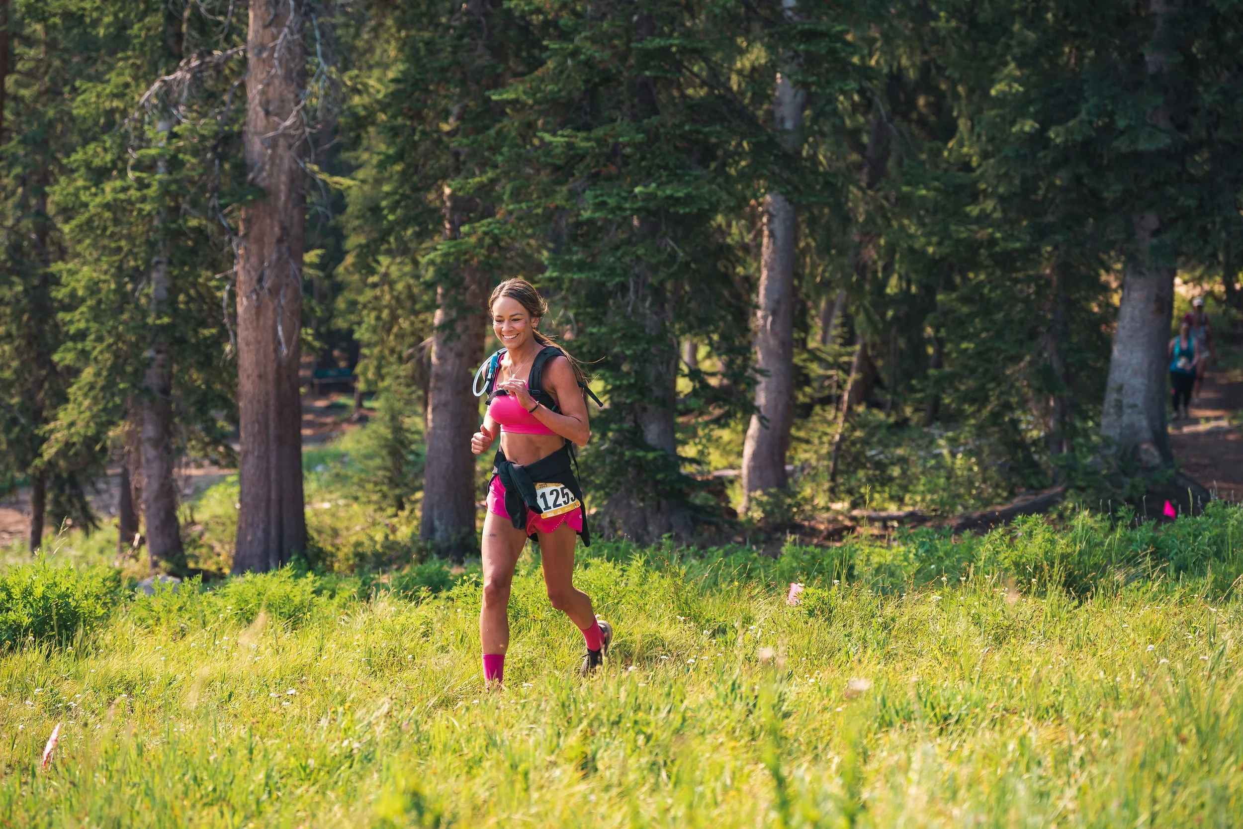 Three women running energetically along a mountain trail, surrounded by lush greenery and scenic views