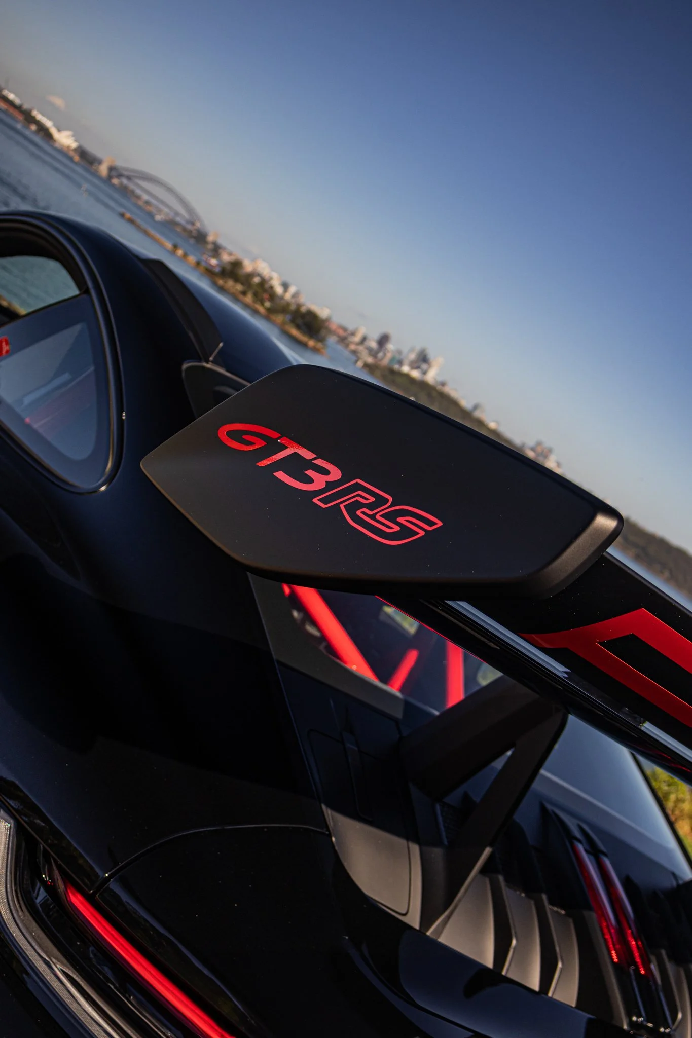 Close-up of a black car with a large rear wing that has red lettering 'GT3 RSR,' with a city skyline and bridge in the background under a clear sky.