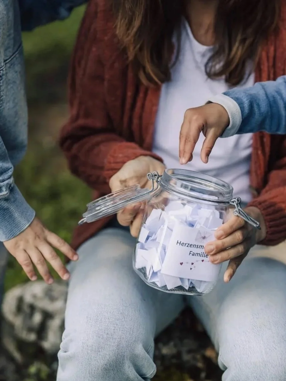 Eine Person hält ein Einmachglas mit kleinen Papierbällen, auf einem ist ein Zettel mit der Aufschrift 'Herzensmutige Familie' und kleinen Herzsymbolen, während eine andere Person ein Papierstück herausnimmt.