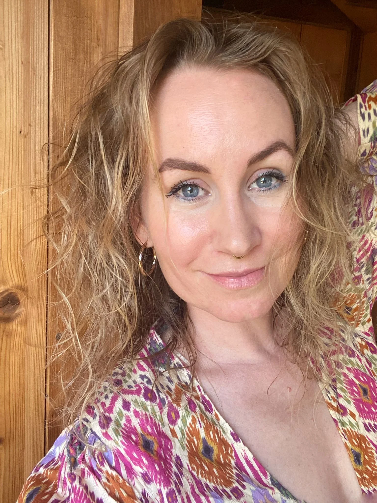 Nahaufnahme einer Frau mit blonden, lockigen Haaren, blauen Augen und dezentem Make-up, trägt eine bunte, gemusterte Bluse, vor einem Holzwand-Hintergrund.