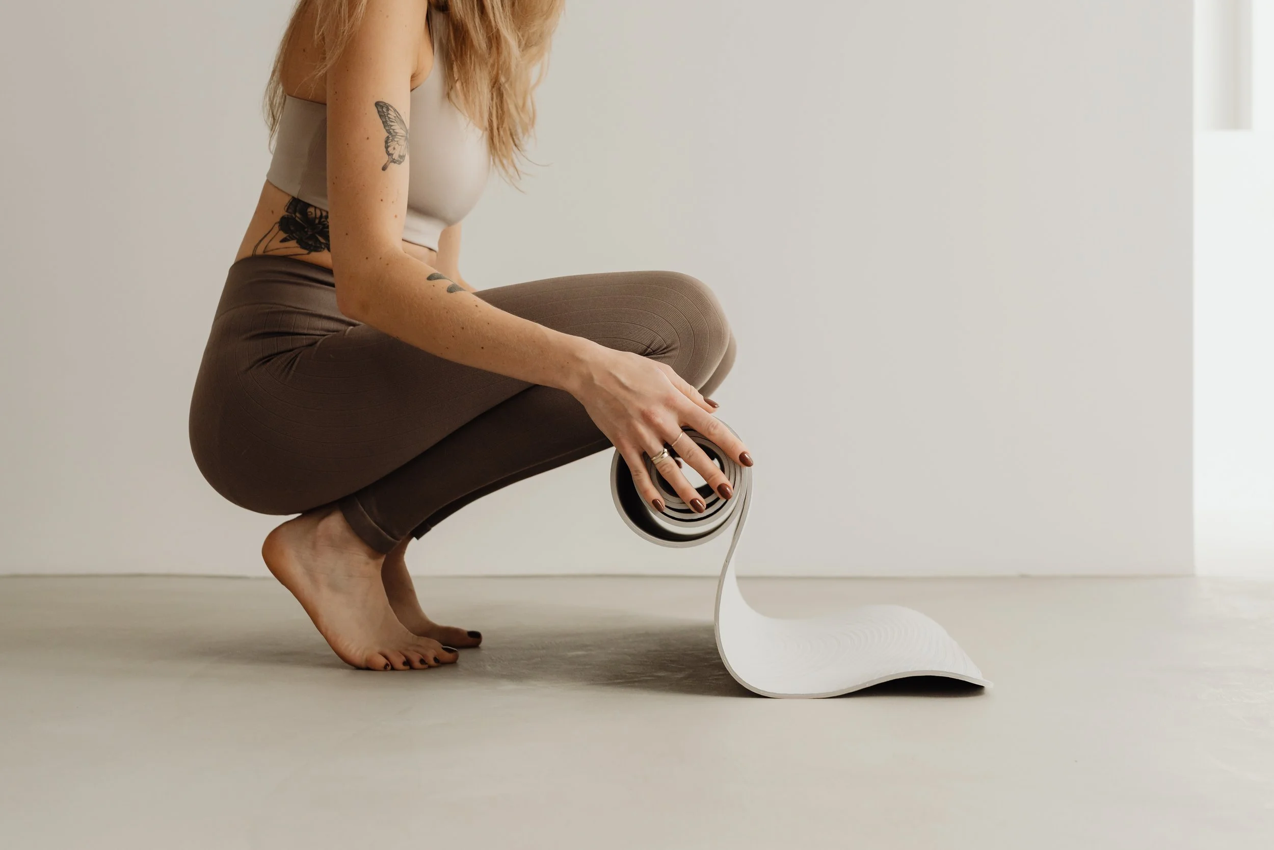Una mujer en posición de yoga realizando una postura de equilibrio, sosteniendo un rodillo de yoga con una mano, con tatuajes visibles en su brazo y costado, vestida con ropa ajustada de color marrón y una camiseta sin mangas de color gris, en un fondo blanco y sin otros objetos visibles.