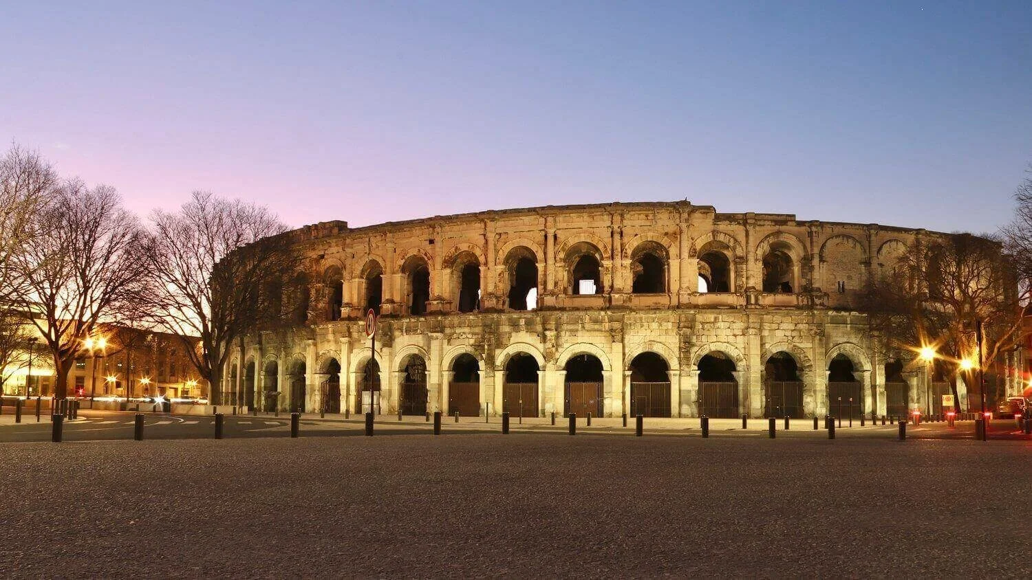 Vue nocturne des Arènes de Nîmes, illuminées contre un ciel crépusculaire. Idéal pour découvrir comment devenir mannequin d’essayage à Nîmes, une ville riche en patrimoine historique romain.
