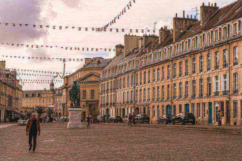 Vue d’une place animée à Caen, avec des bâtiments typiques bordés de drapeaux et une statue centrale. Idéal pour découvrir comment devenir mannequin d’essayage à Caen, une ville où l’histoire et la modernité coexistent.