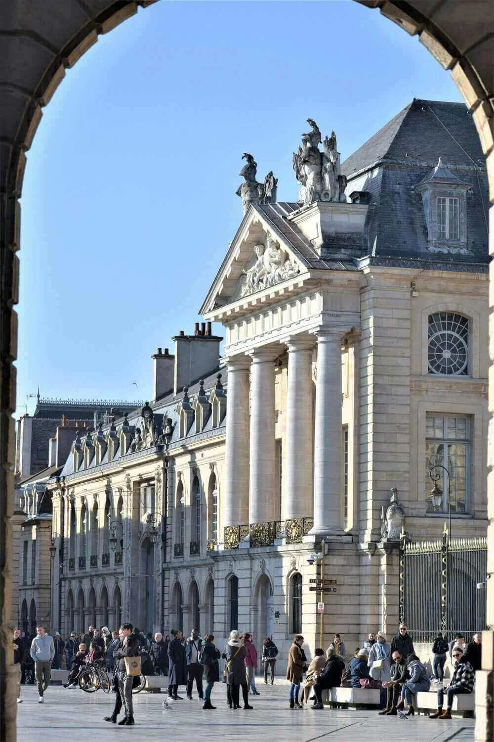 Vue d’une place animée à Dijon, ornée d’un monument historique imposant avec des colonnes et des sculptures décoratives. Idéal pour découvrir comment devenir mannequin d’essayage dans une ville riche en patrimoine architectural.