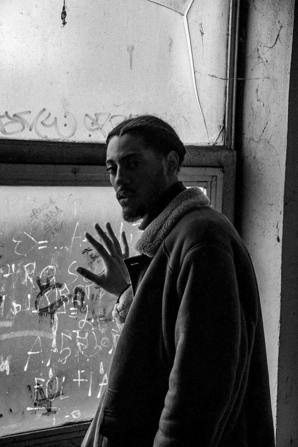 Vue en noir et blanc d’un homme debout devant une fenêtre recouverte de graffitis, dans un environnement urbain à Montreuil. Idéal pour découvrir comment devenir mannequin d’essayage à Montreuil, une ville où l’art et la créativité se rencontrent.